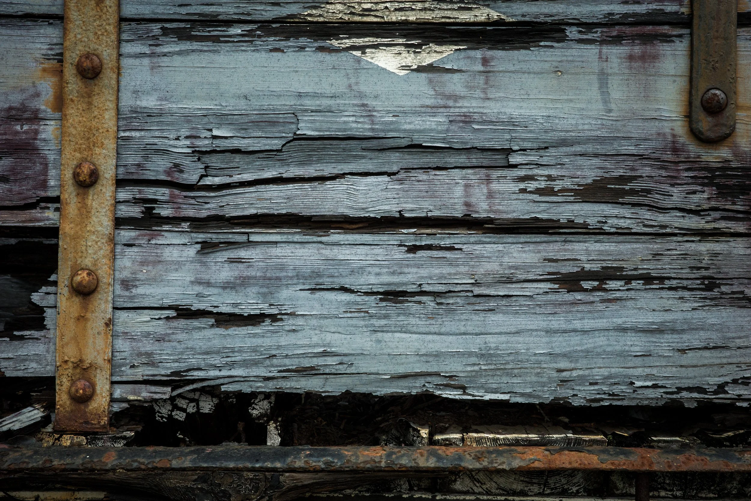 Close-up of an old, weathered wooden surface with peeling paint, rusted metal strips, and rusty bolts.