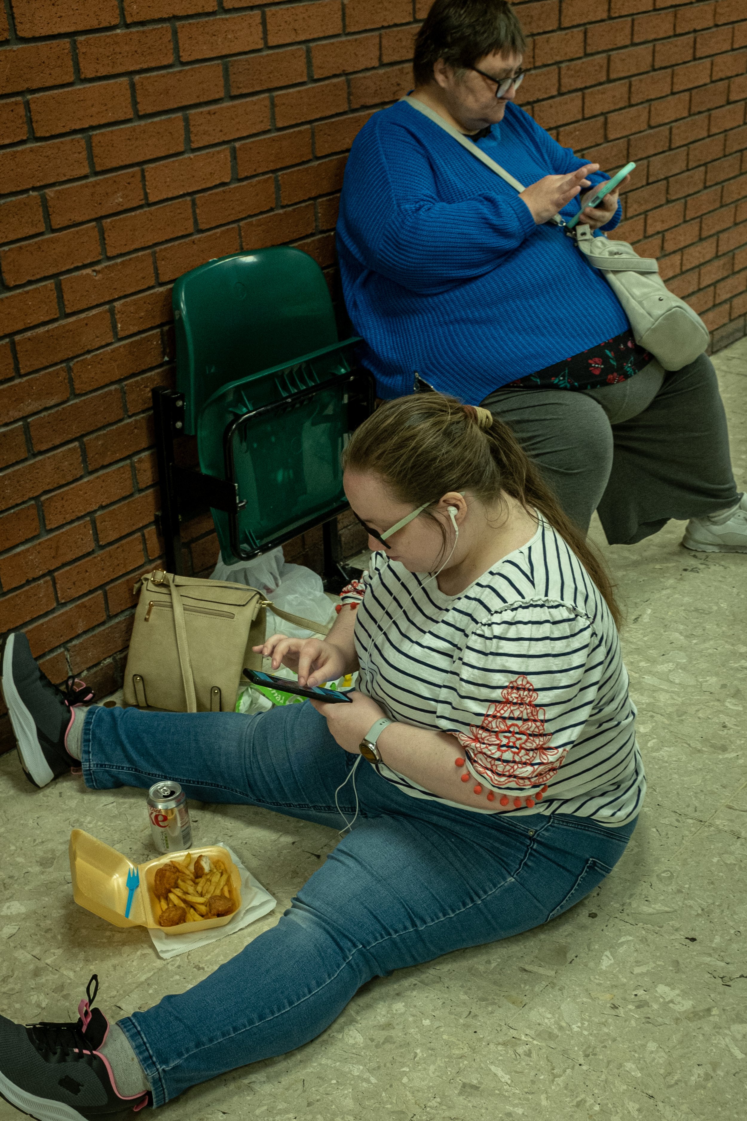 A woman with a striped shirt and glasses sitting on the floor, playing with her phone, with a food container and a can nearby. A woman in a blue sweater and glasses is sitting on a chair against a brick wall, looking at her phone. There are chairs st