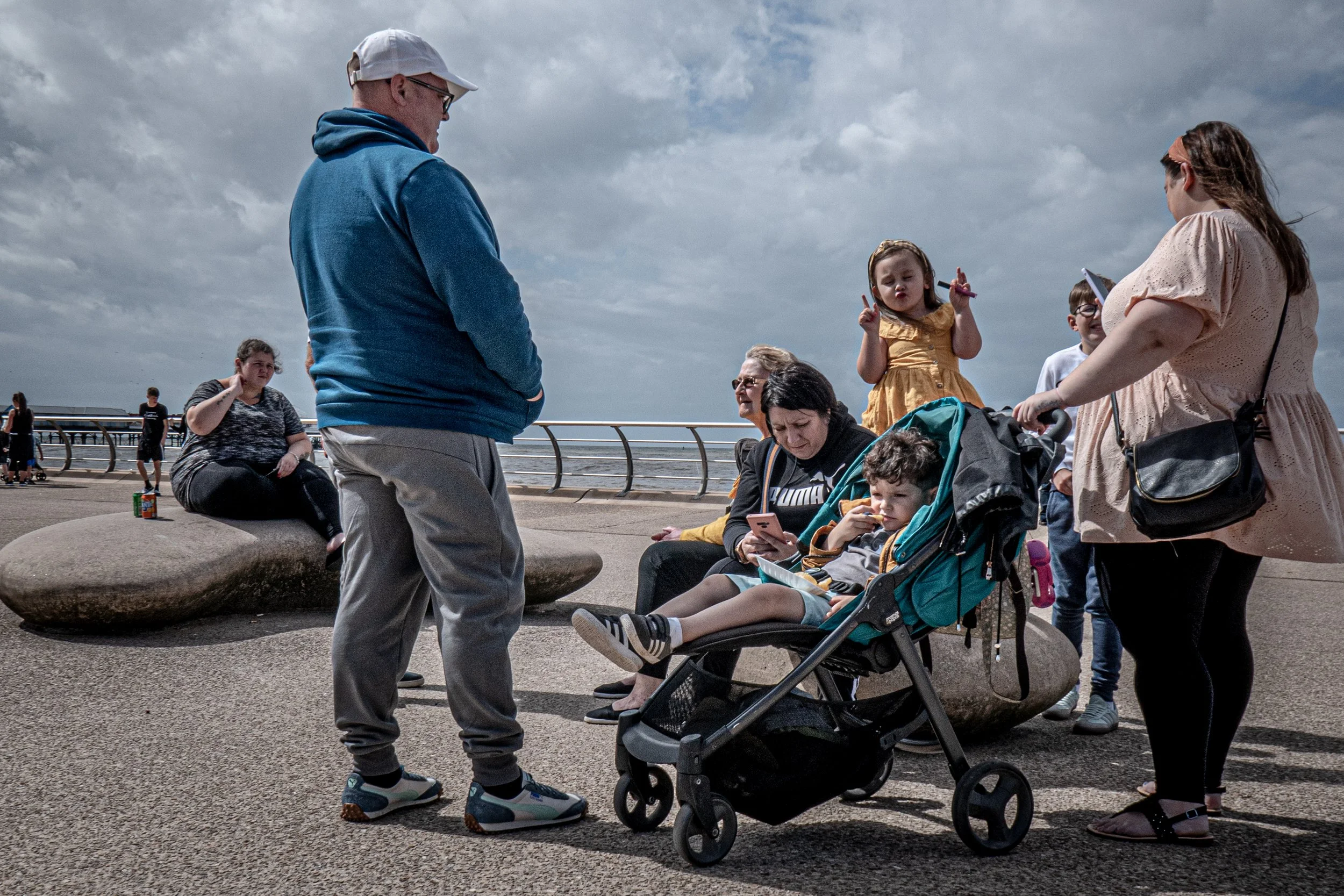 Group of people talking outdoors near the ocean on a cloudy day. One woman is sitting on a large rock, while others are standing or sitting nearby, including a girl in a yellow dress and a woman with a stroller.