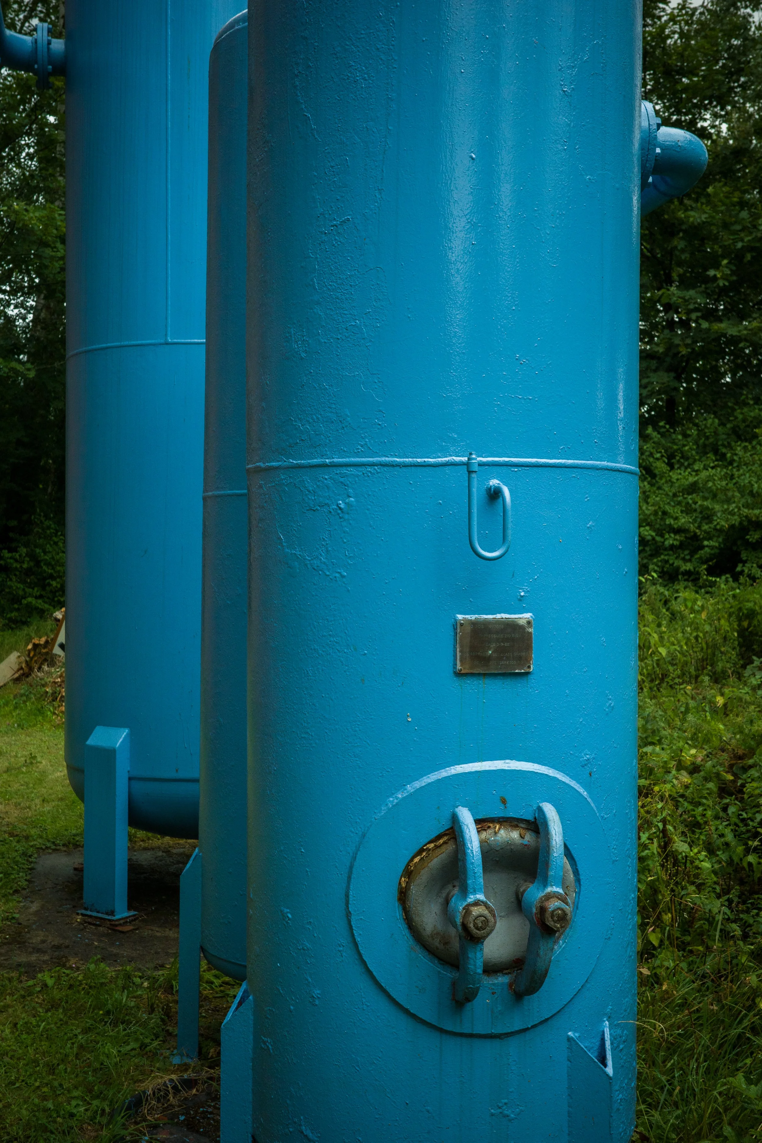 Close-up of large blue industrial water boiler with pipes and a metal plaque, set outdoors with trees in the background.