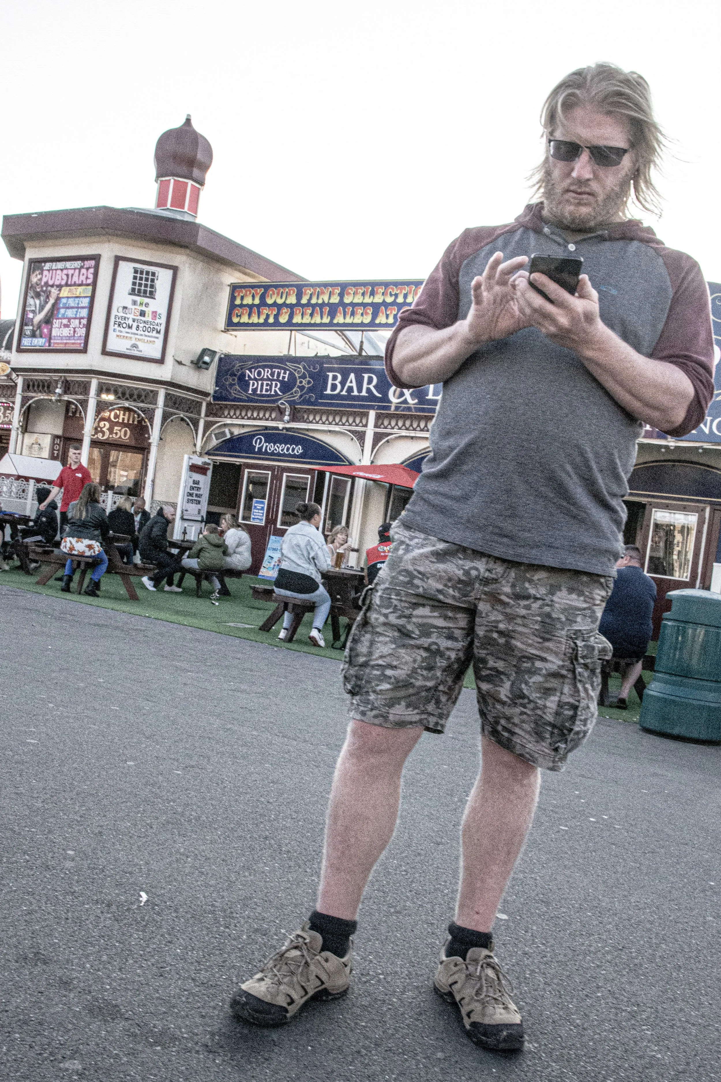 A man with long blond hair, wearing sunglasses, a gray and maroon hoodie, camouflage shorts, and hiking shoes, is standing outdoors using a smartphone. In the background, there is a busy outdoor area with people sitting and standing, and a building w