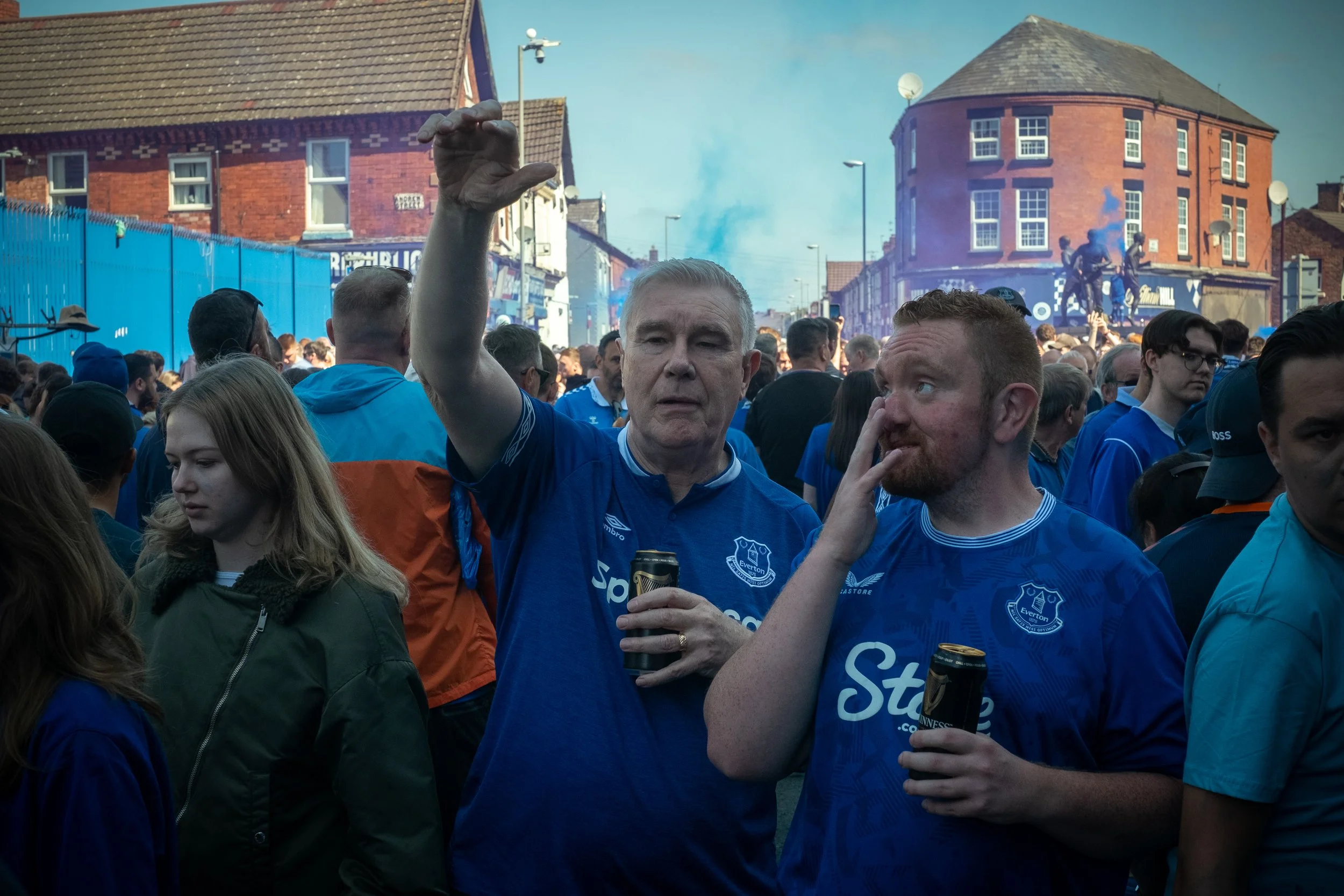 Crowd of people wearing blue, celebrating outside on the street with some holding drinks, in front of buildings and a corner in a city.