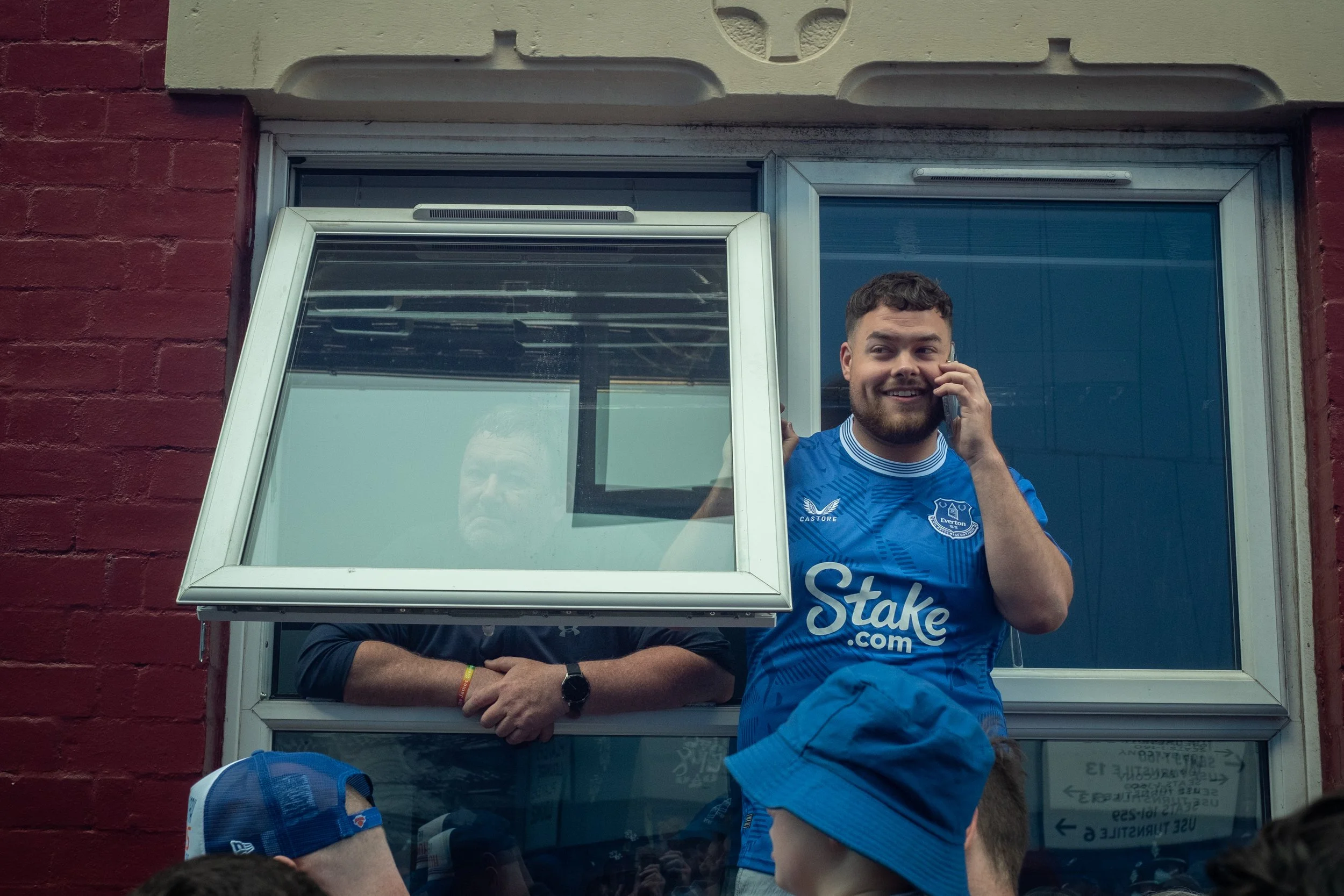 A man in a blue football jersey talking on a cell phone and smiling, seen through an open window, with other people below wearing blue caps.
