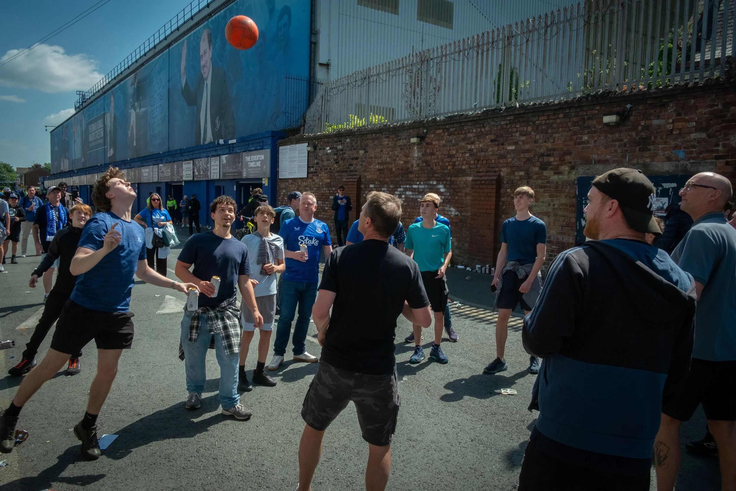 Group of young males watching a basketball bounce near a brick wall and metal fence at an outdoor event on a sunny day
