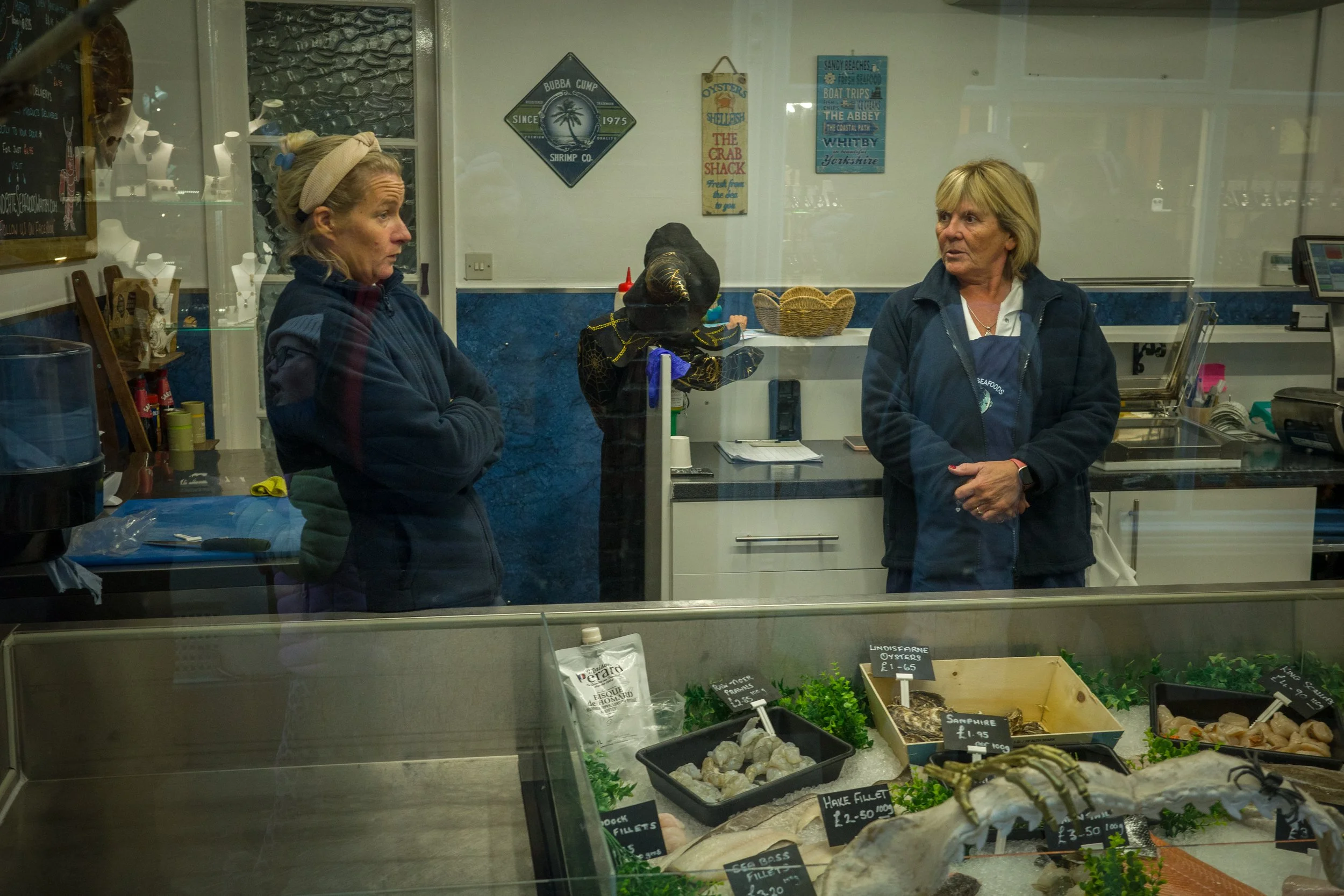 Two women having a conversation inside a seafood shop, seen through a glass display case with various fresh seafood and prices labeled.