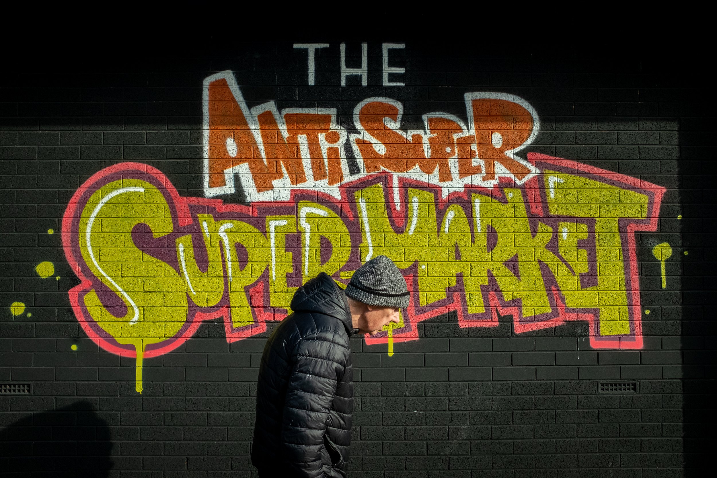 A person wearing a gray beanie and black puffer jacket looking down in front of a colorful graffiti wall that reads 'The Artsy Summer Spectrum' with vibrant pink, yellow, and orange lettering.