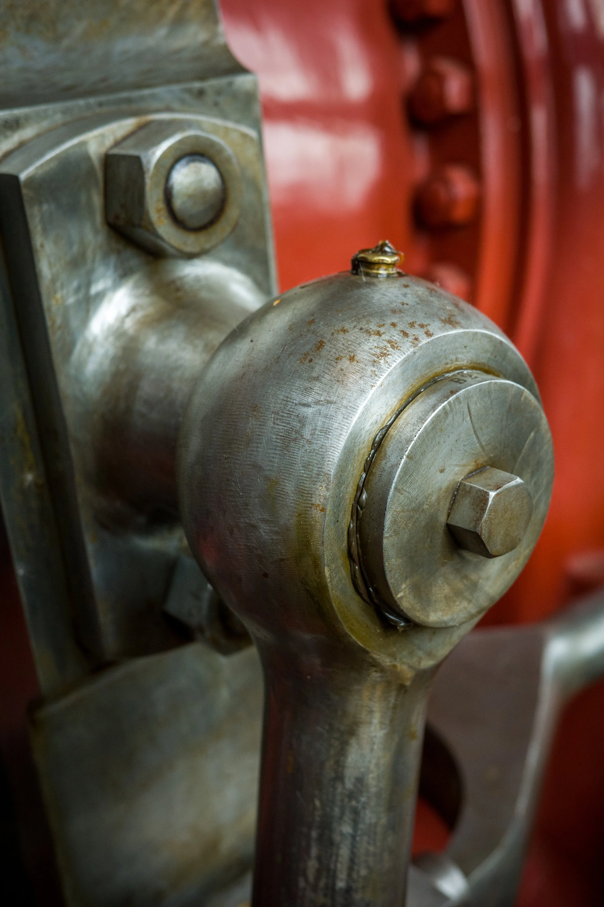 A close-up of a metallic valve with rust spots attached to a pipe, with a chain hanging from the valve, and a red background with large bolts.