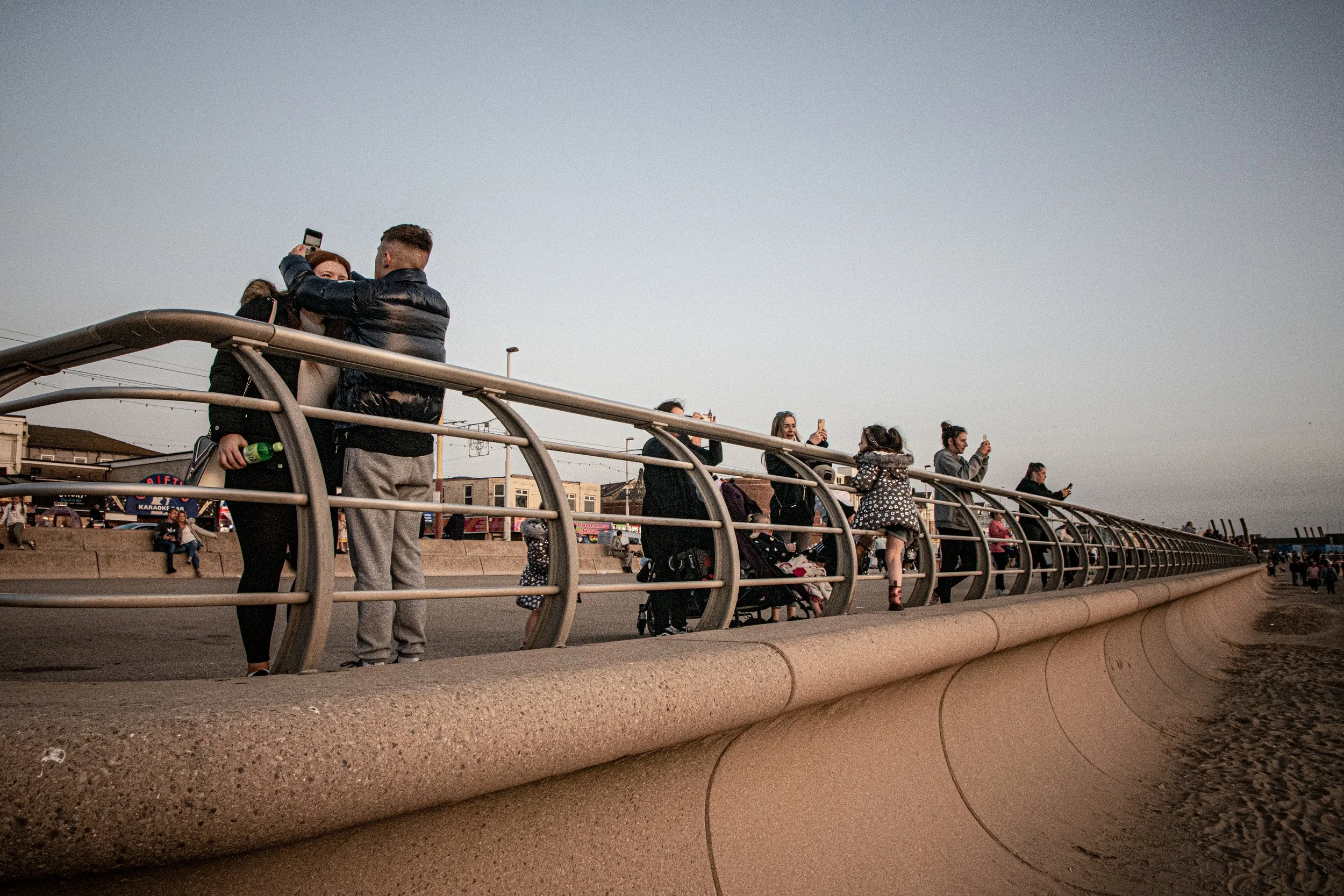People standing on a bridge railing, some taking photos, others socializing, under a clear sky.