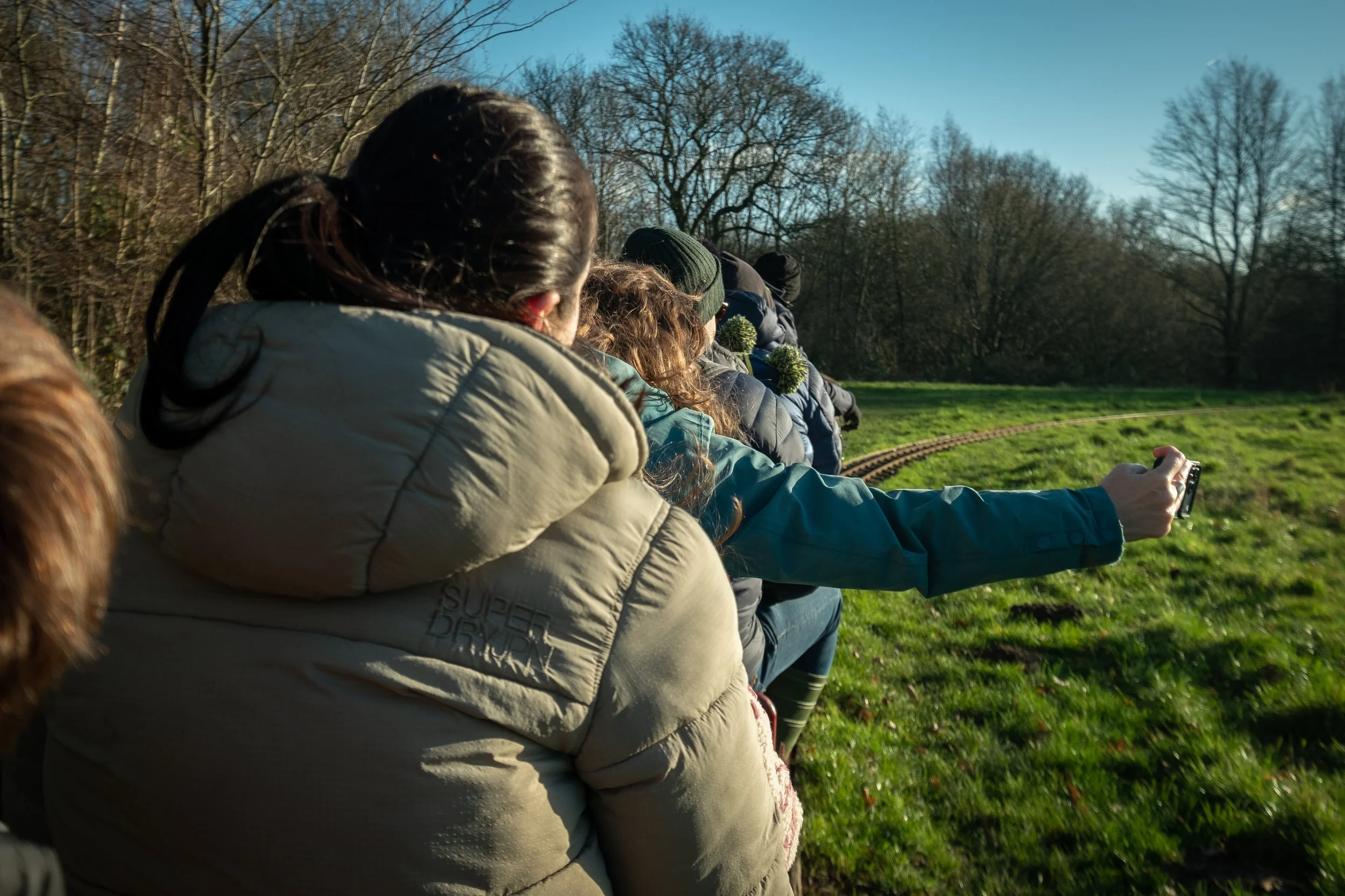 A row of people with jackets and hats sitting outdoors in a park or open field, some holding or taking pictures with a phone, with leafless trees and a clear blue sky in the background.