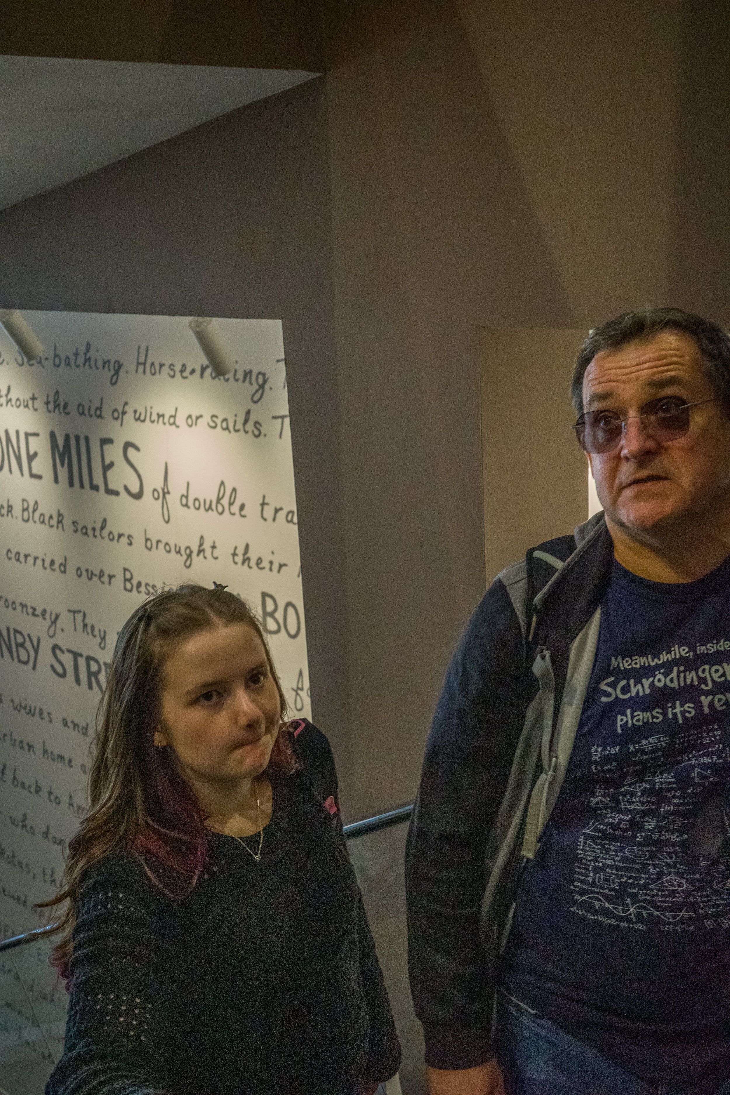 A young girl and a man standing near a staircase with a handwritten poster in the background. The girl has long hair with purple highlights, and the man is wearing glasses and a t-shirt with a scientific diagram.