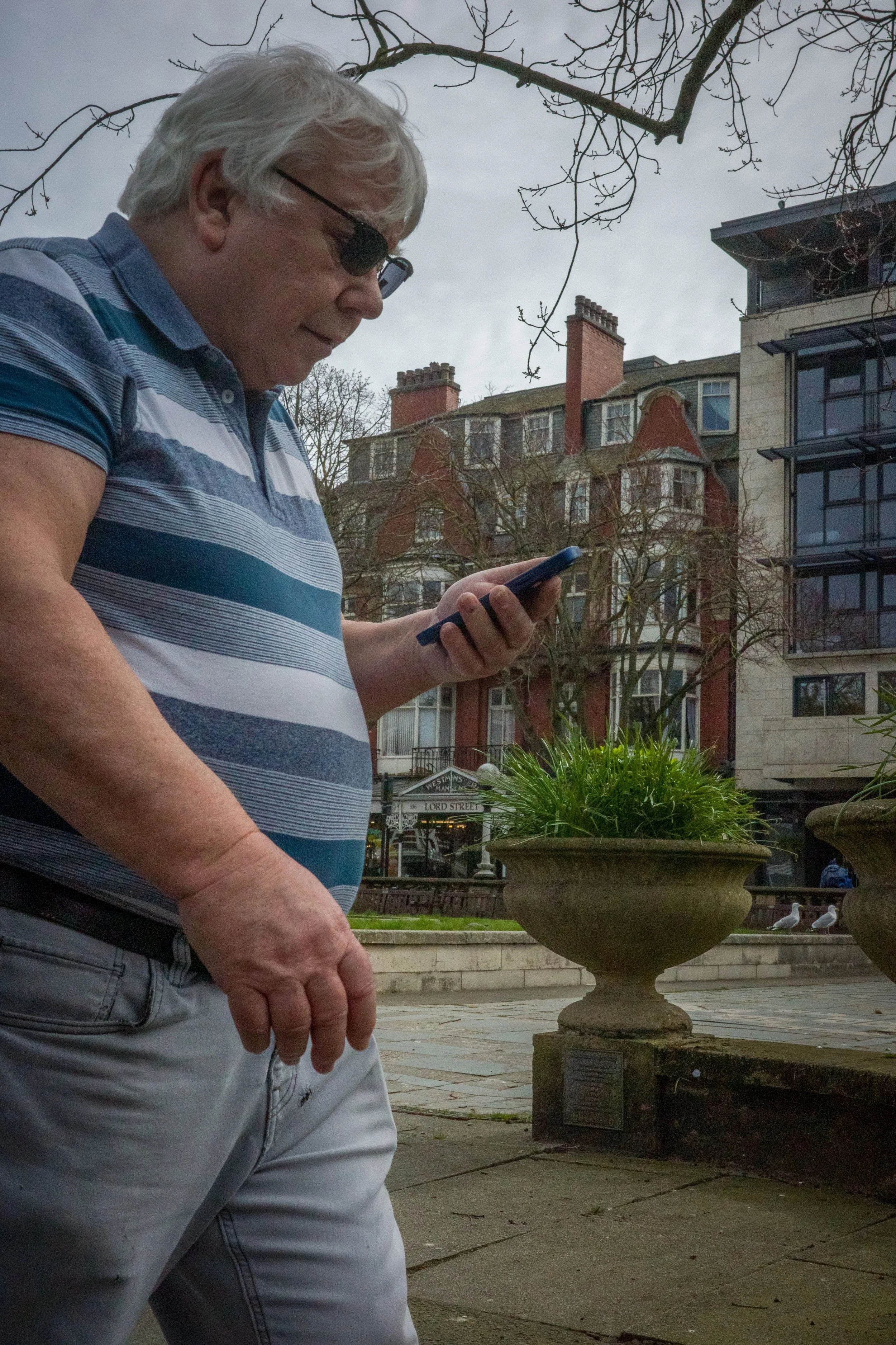 A man wearing glasses and a striped blue and white polo shirt is looking at his phone, standing outdoors in an urban setting with a large flowerpot and multiple residential buildings in the background.