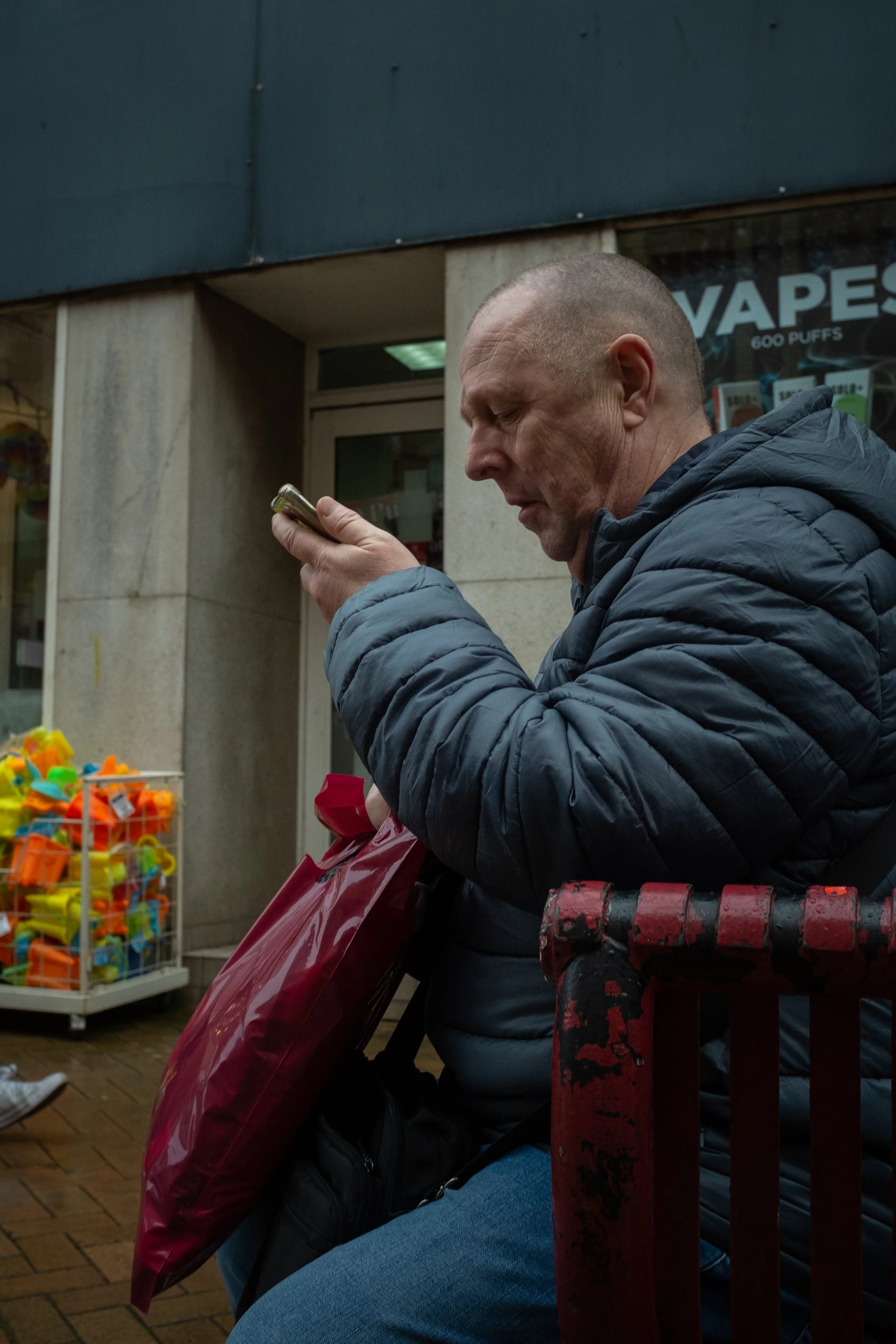 A man in a blue jacket is sitting on a bench, looking at his phone, with a bag on his lap. In the background, there is a store window with a sign that reads 'Vapes'. There is a display with colorful toys or small items on the left side.
