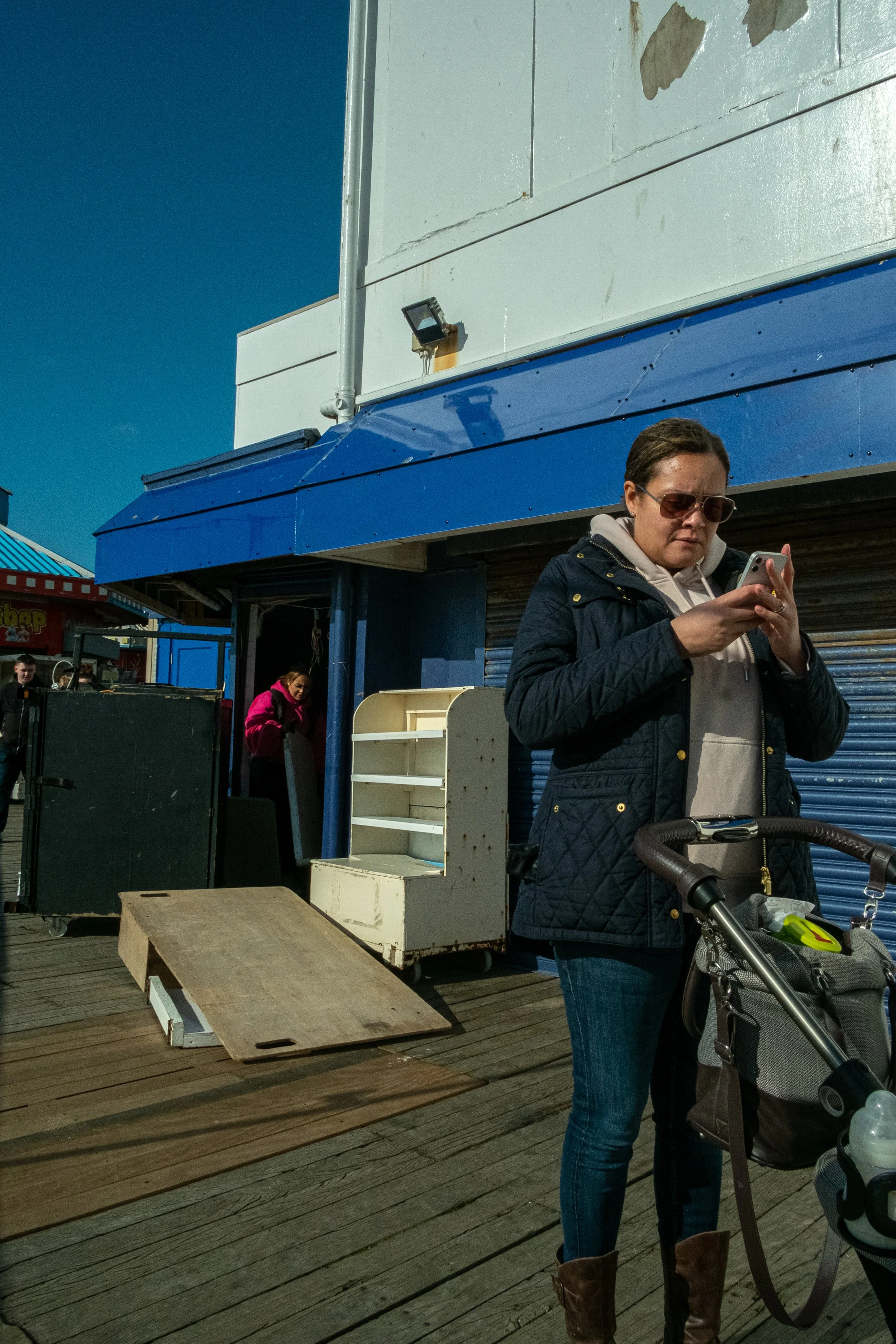 A woman wearing a dark quilted jacket and sunglasses standing outdoors on a wooden deck next to a stroller, with a person in the background. There is a white display shelf and an open wooden ramp on the ground. The background includes a white buildin