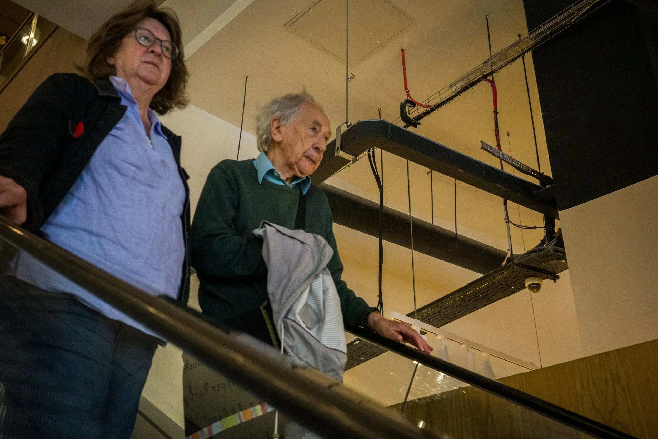 Two women on an indoor balcony, looking down. One woman with glasses and dark hair is on the left, and the other elderly woman with gray hair is holding a jacket in her hand.