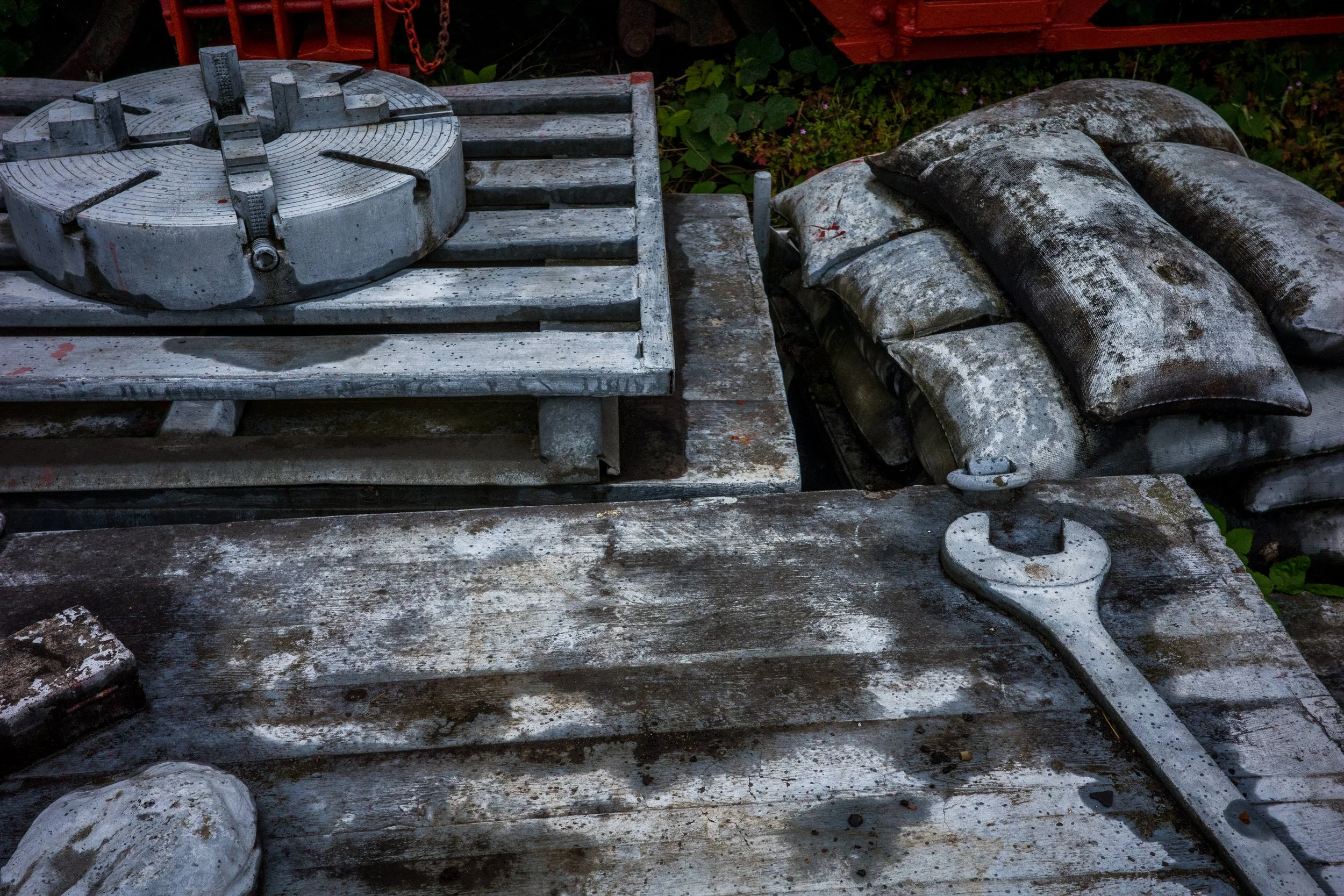 Outdoor scene with old weathered wooden tables, bags, and a metal object with a circular base, surrounded by plants and a red piece of equipment in the background.