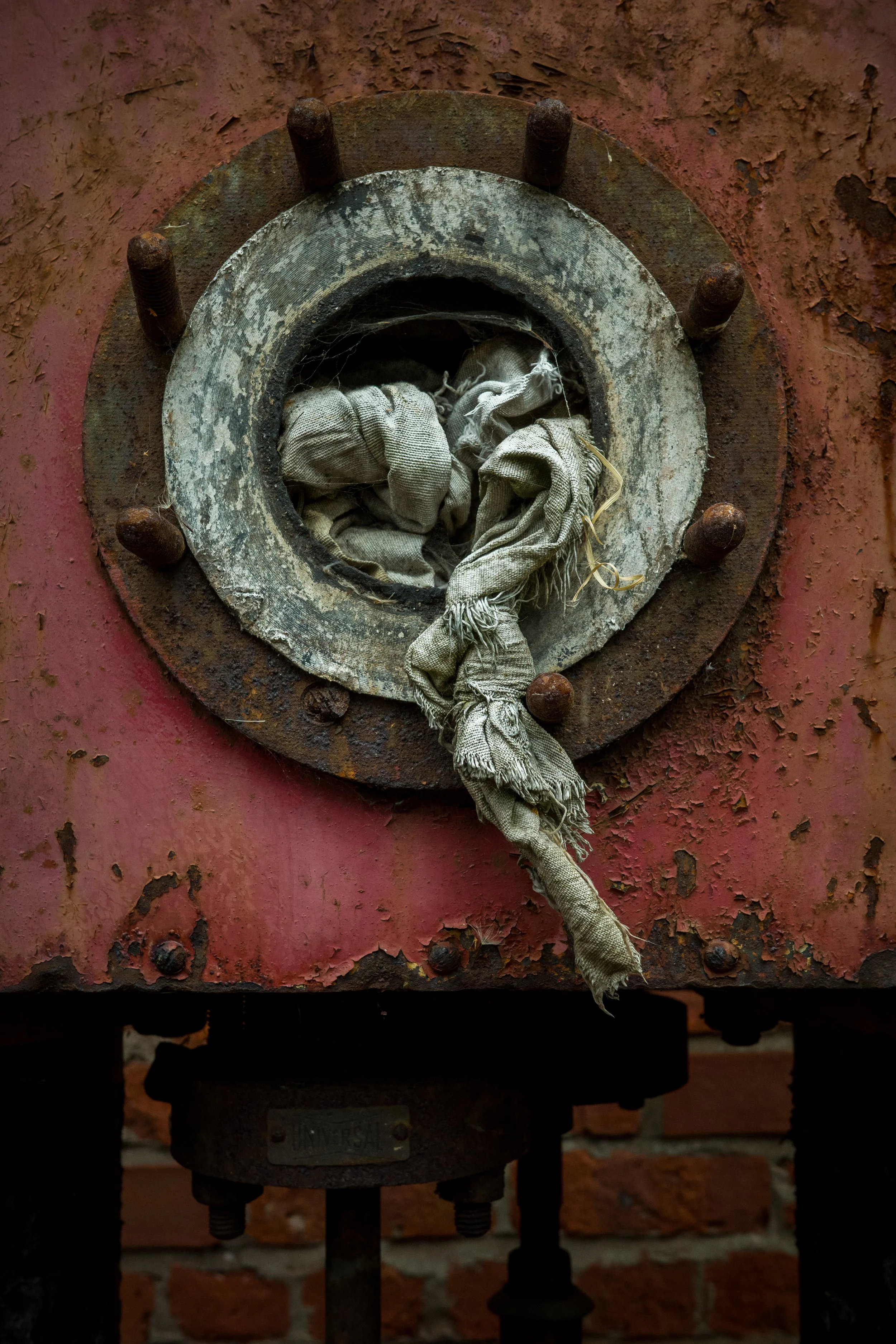Close-up of a rusty, worn industrial machine or equipment part with a fabric rag tangled around it, mounted on a red, weathered brick wall.
