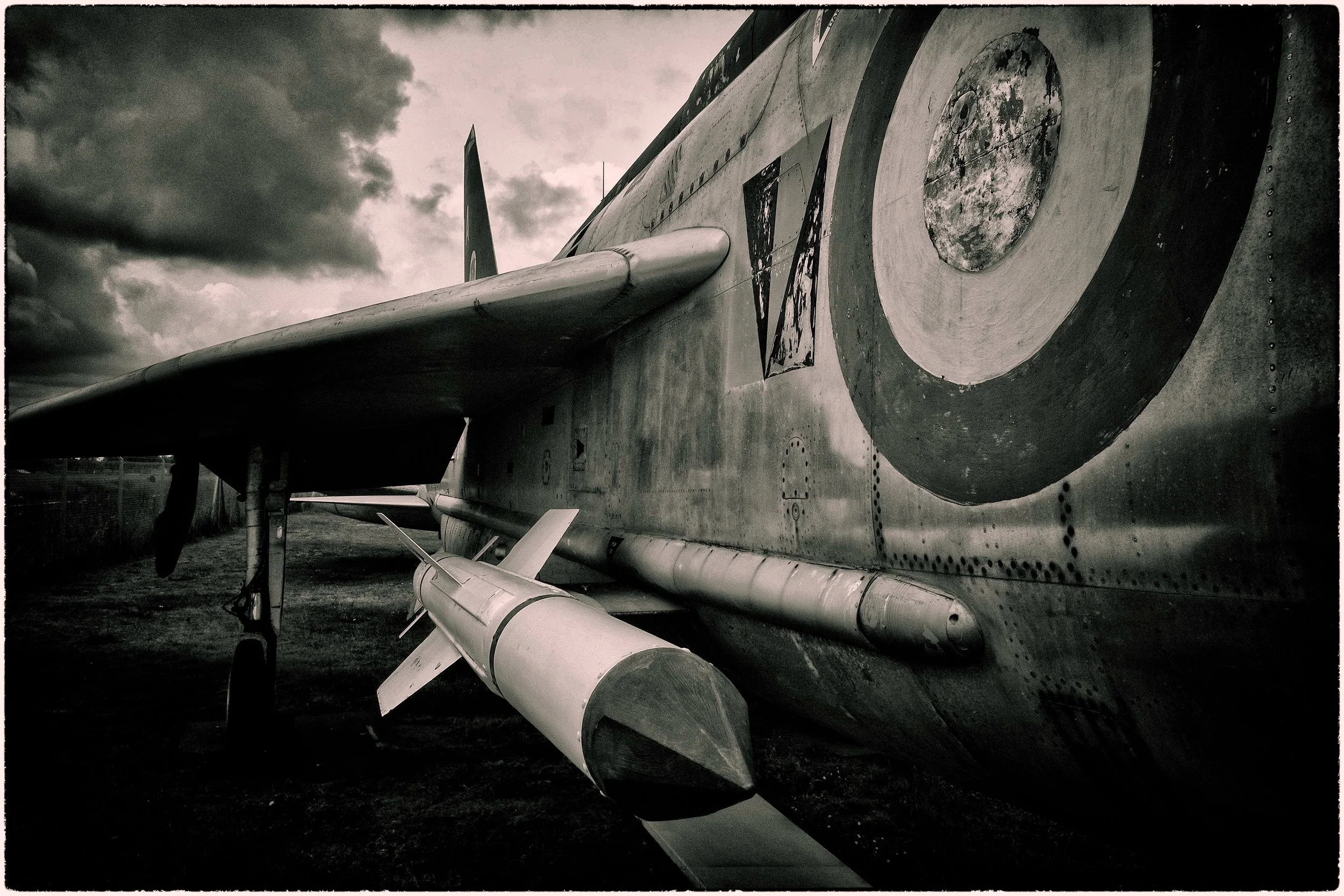 Close-up of a retired military fighter jet aircraft exterior showing its wing, missile, and insignia under cloudy sky.