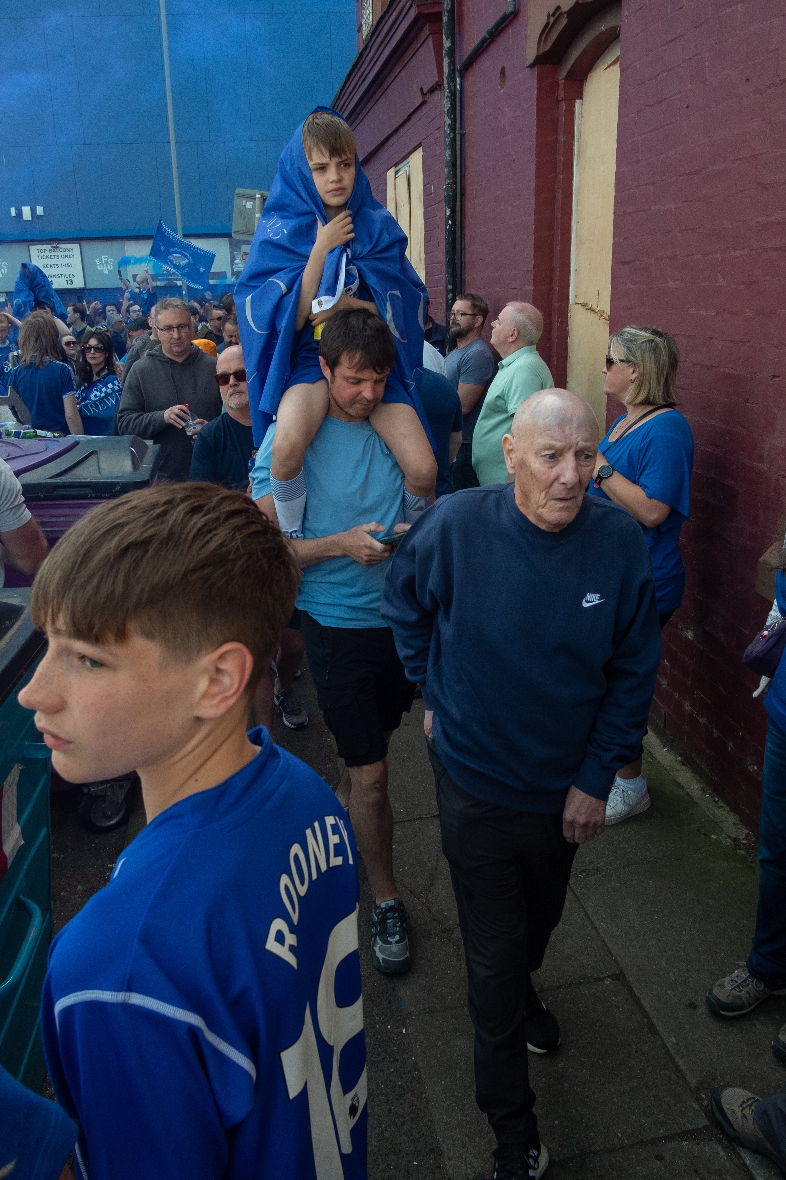 Crowd of sports fans, including children and adults, gathering outdoors, some wearing blue shirts, with one young boy sitting on an older man's shoulders wrapped in a blue caped jacket.