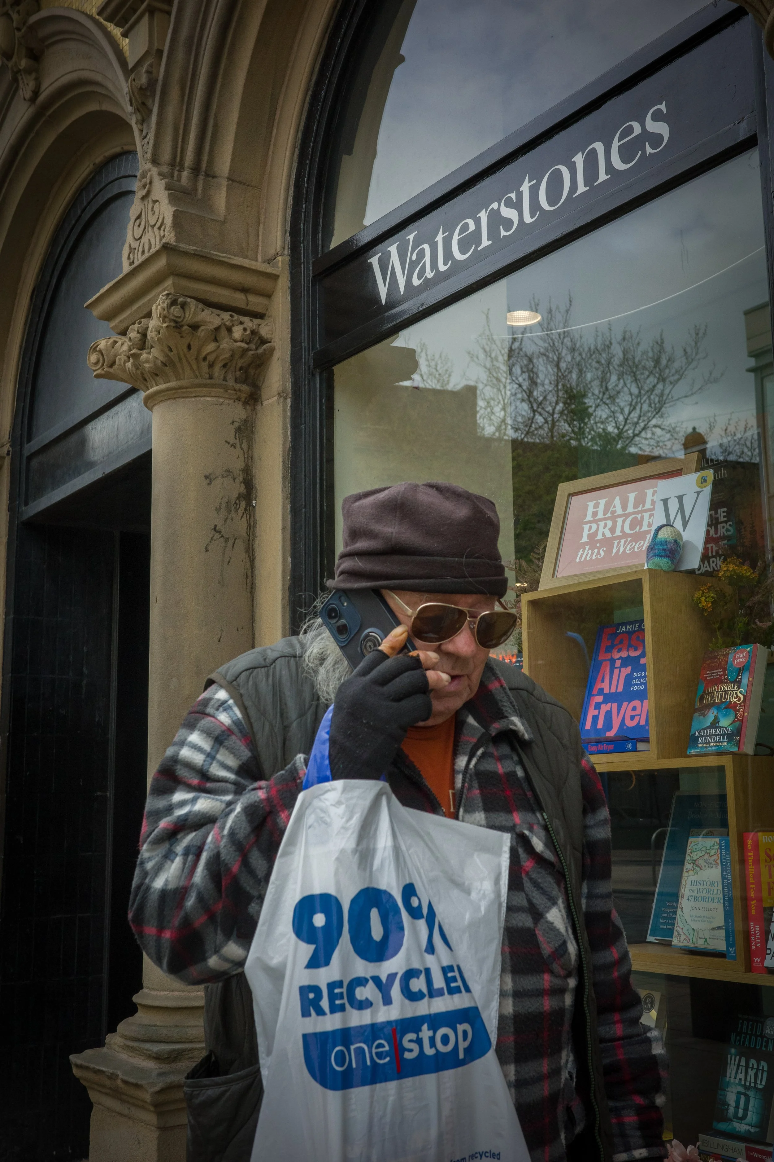 A woman wearing sunglasses, a brown hat, and a plaid jacket is holding a plastic bag that says "Recycle" and is talking on a cell phone while standing outside a bookstore with a sign for a book sale.