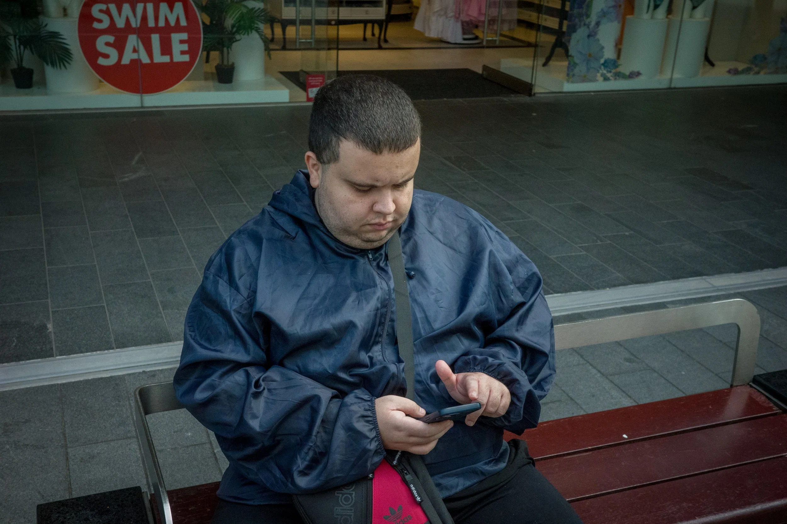 A young man is sitting on a park bench, looking down at his smartphone. He is wearing a dark blue jacket and has short, dark hair. Behind him is a store with a red sign that reads 'SWIM SALE' in a shopping mall or outdoor shopping area.