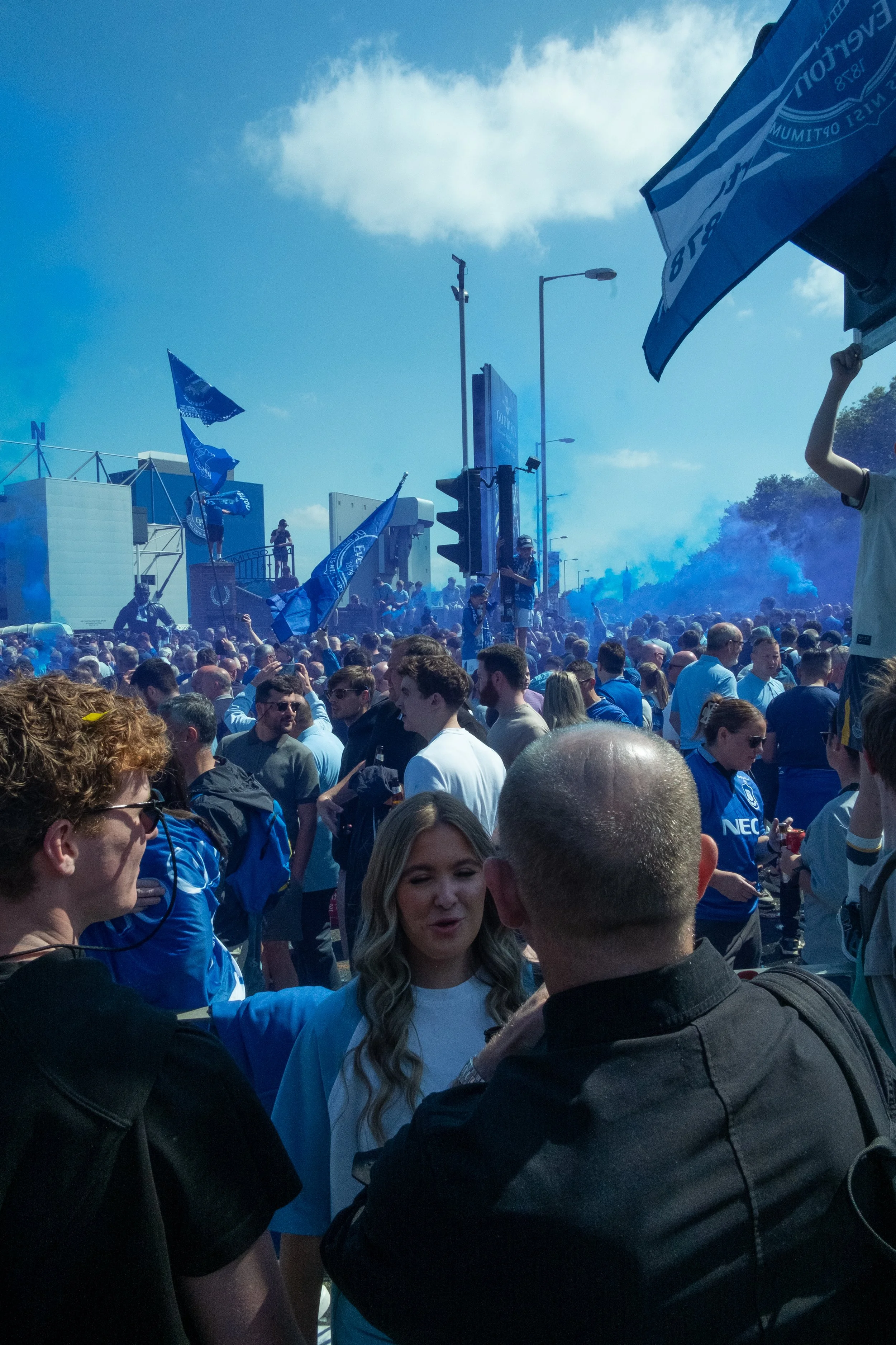 Crowd of football fans gathered outdoors, some wearing team jerseys, waving flags, and holding blue smoke flares during a sunny day.