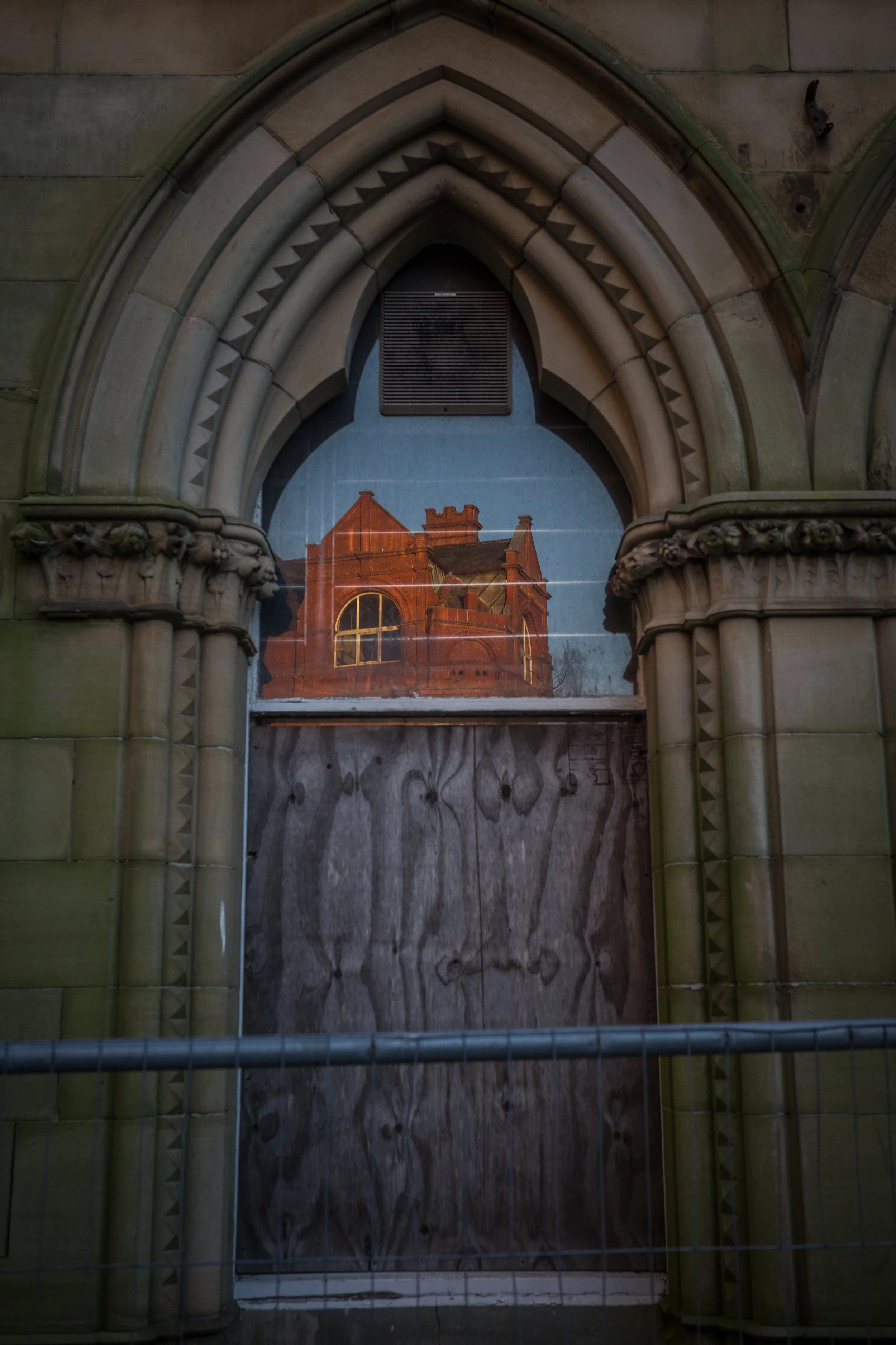 Reflection of a red brick Victorian-style house with a large arched window, seen through a small window of an old building with ornate stonework and construction fencing.