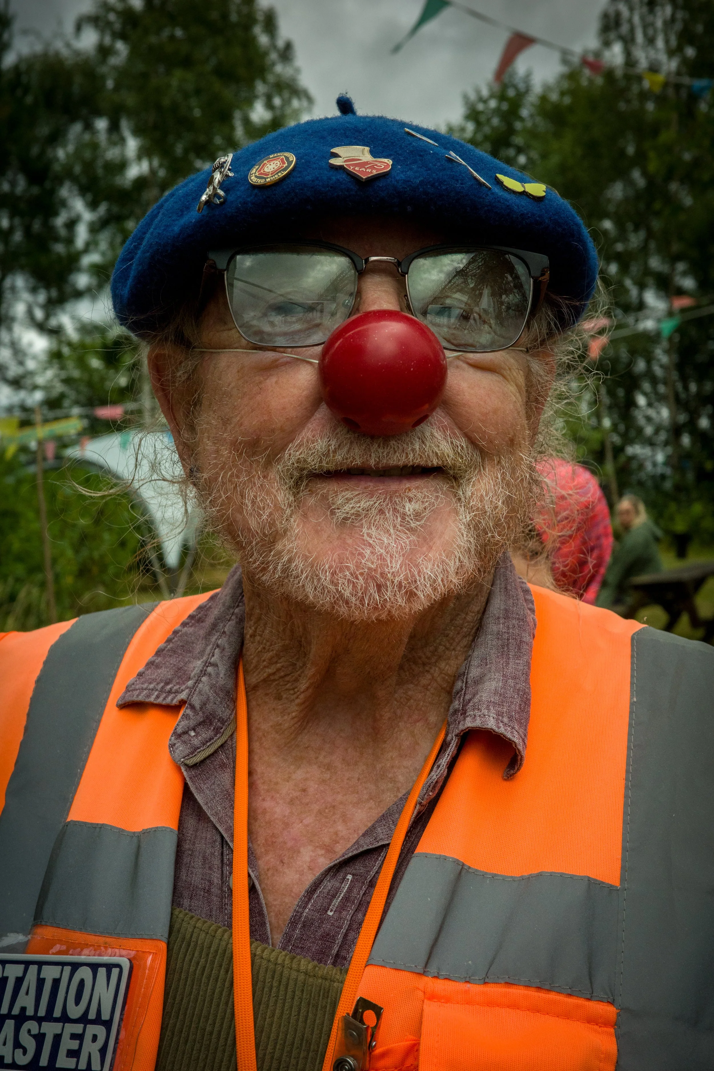 A man wearing glasses, a clown nose, a blue hat decorated with pins, and an orange safety vest. There are trees and tents in the background.