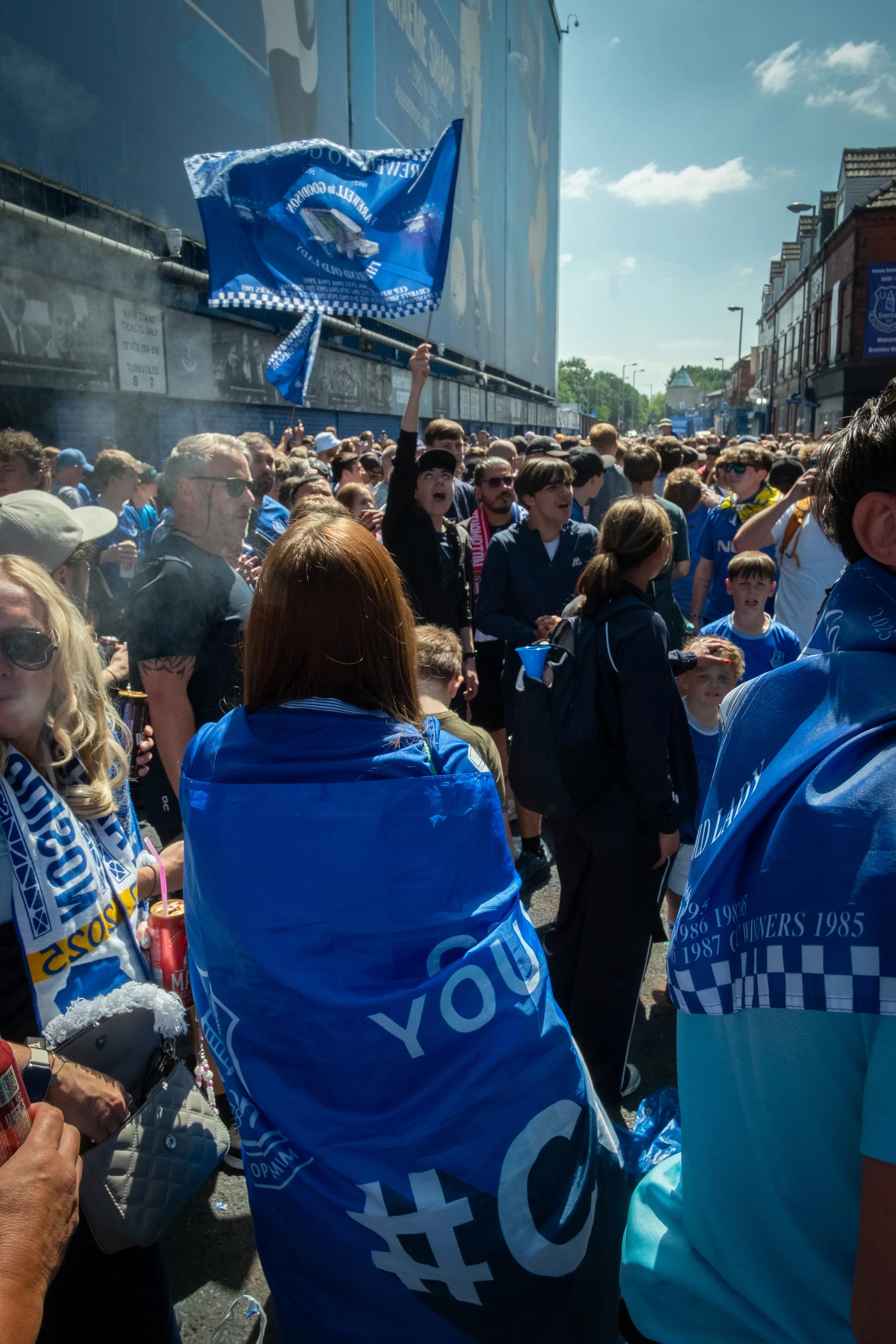 Crowd of football fans dressed in blue, some with scarves and flags, gathered outdoors on a sunny day for a sporting event.