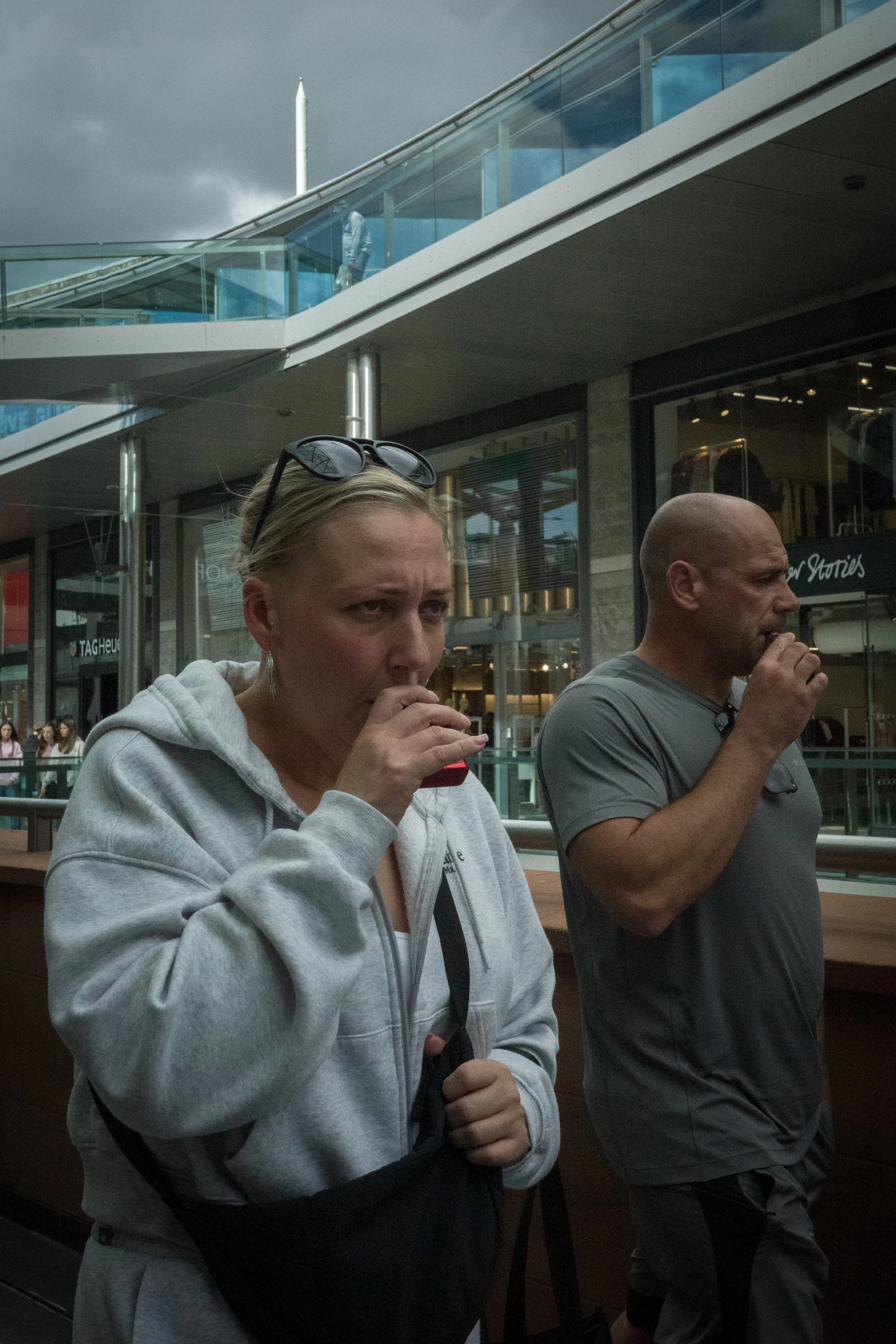 A woman and a man standing outdoors, drinking from cans, with a modern building and a glass staircase in the background.