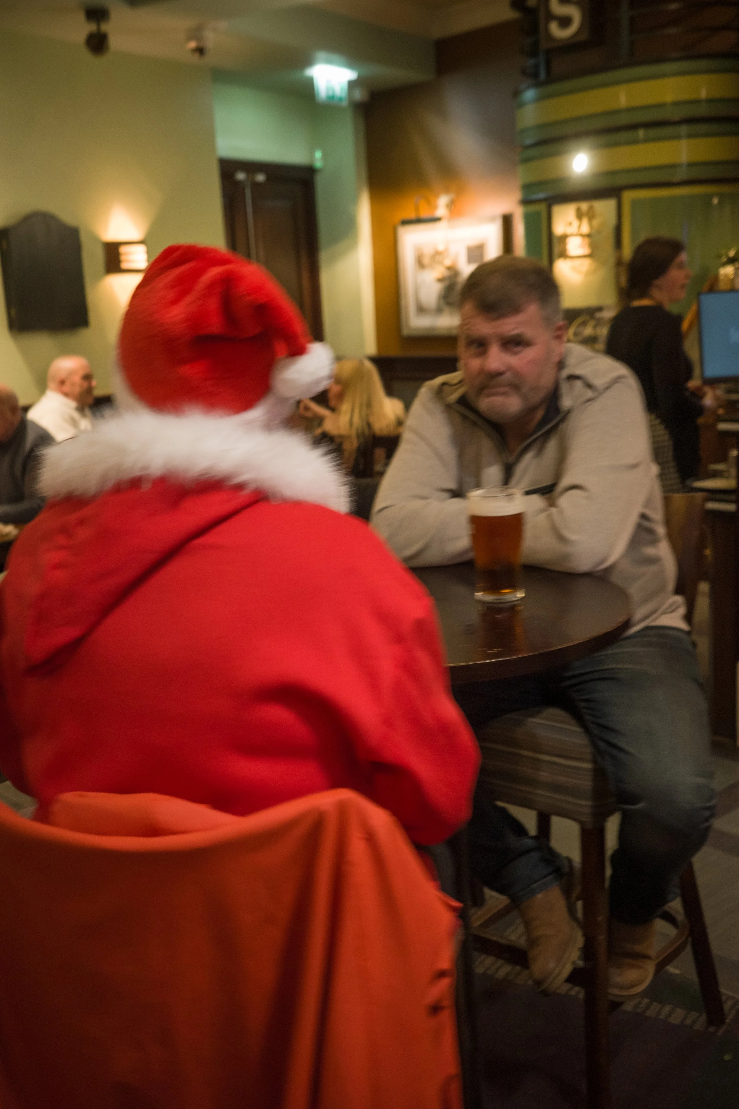 A man sitting at a round table in a pub, facing a person dressed as Santa Claus with a red hat and red coat, in a festive setting with warm lighting and other patrons in the background.