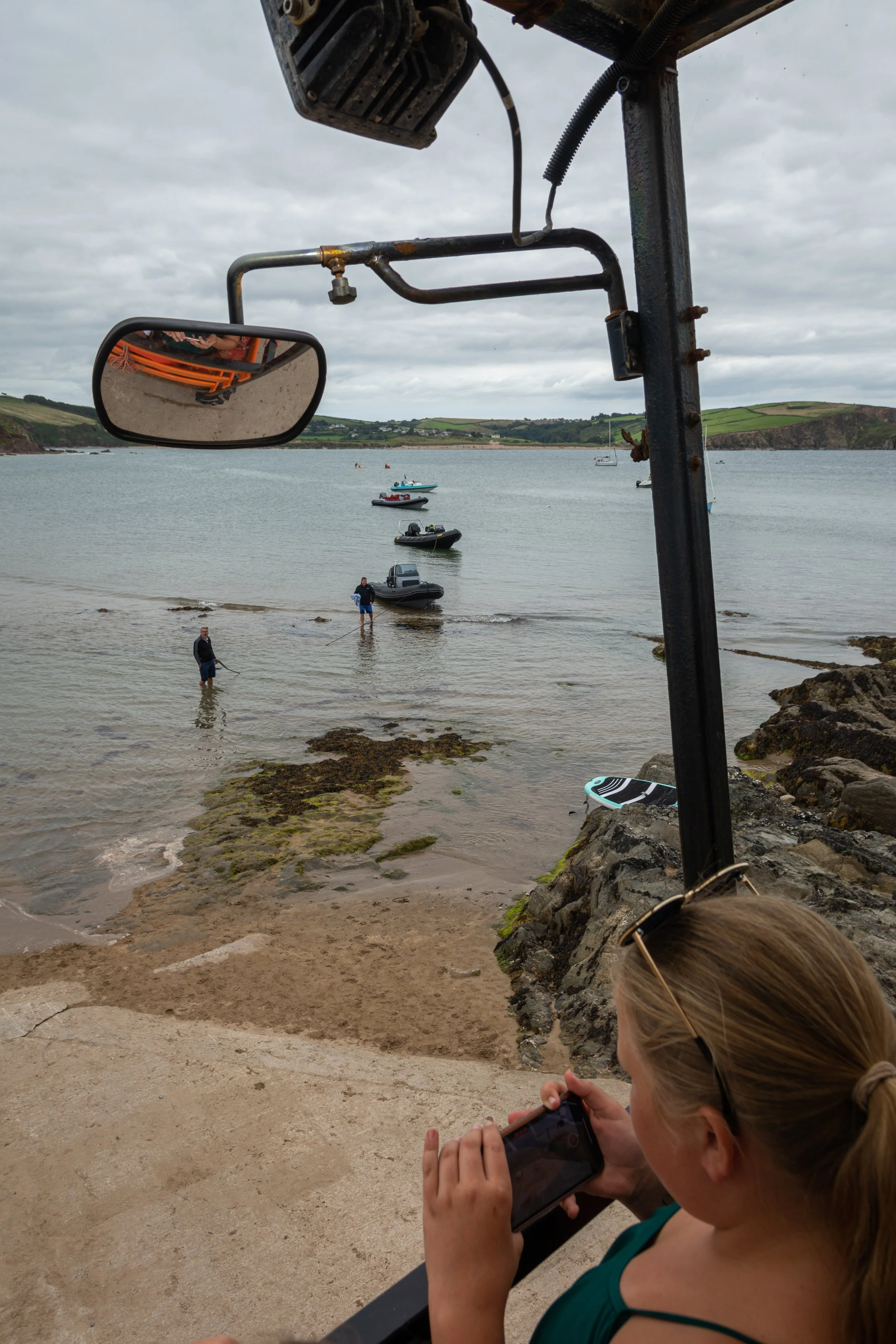 A woman with blonde hair, wearing sunglasses and a teal top, sitting on a rock at the beach, using her phone. In the background, there are some boats in the water and two people standing near the shoreline. A mirror reflects part of an orange-colored