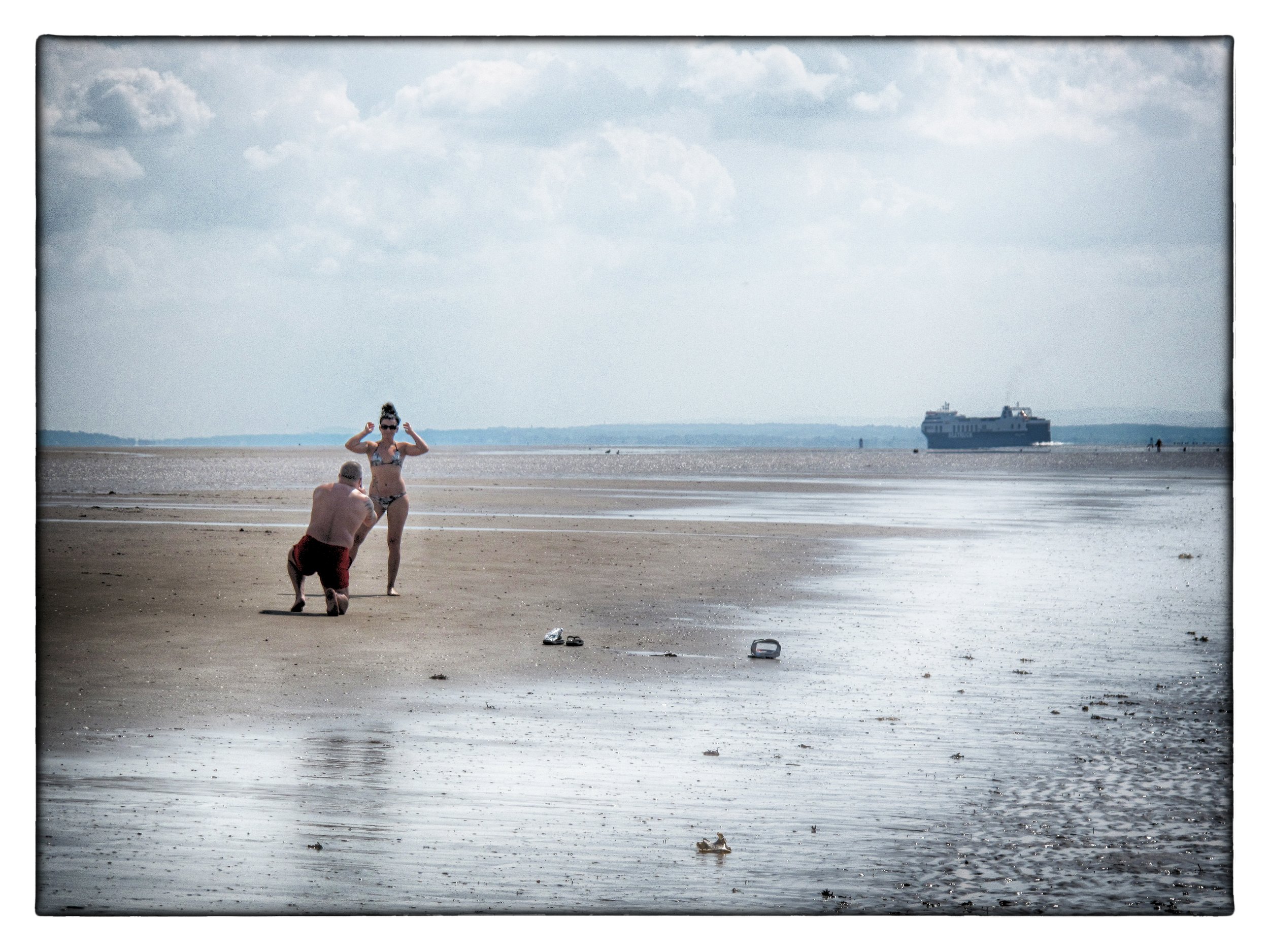 A man is kneeling in front of a woman on a beach, taking a photo of her, while she stands in a bikini with her arms raised. The beach is mostly empty, with a ship in the distance and some scattered objects on the sand.