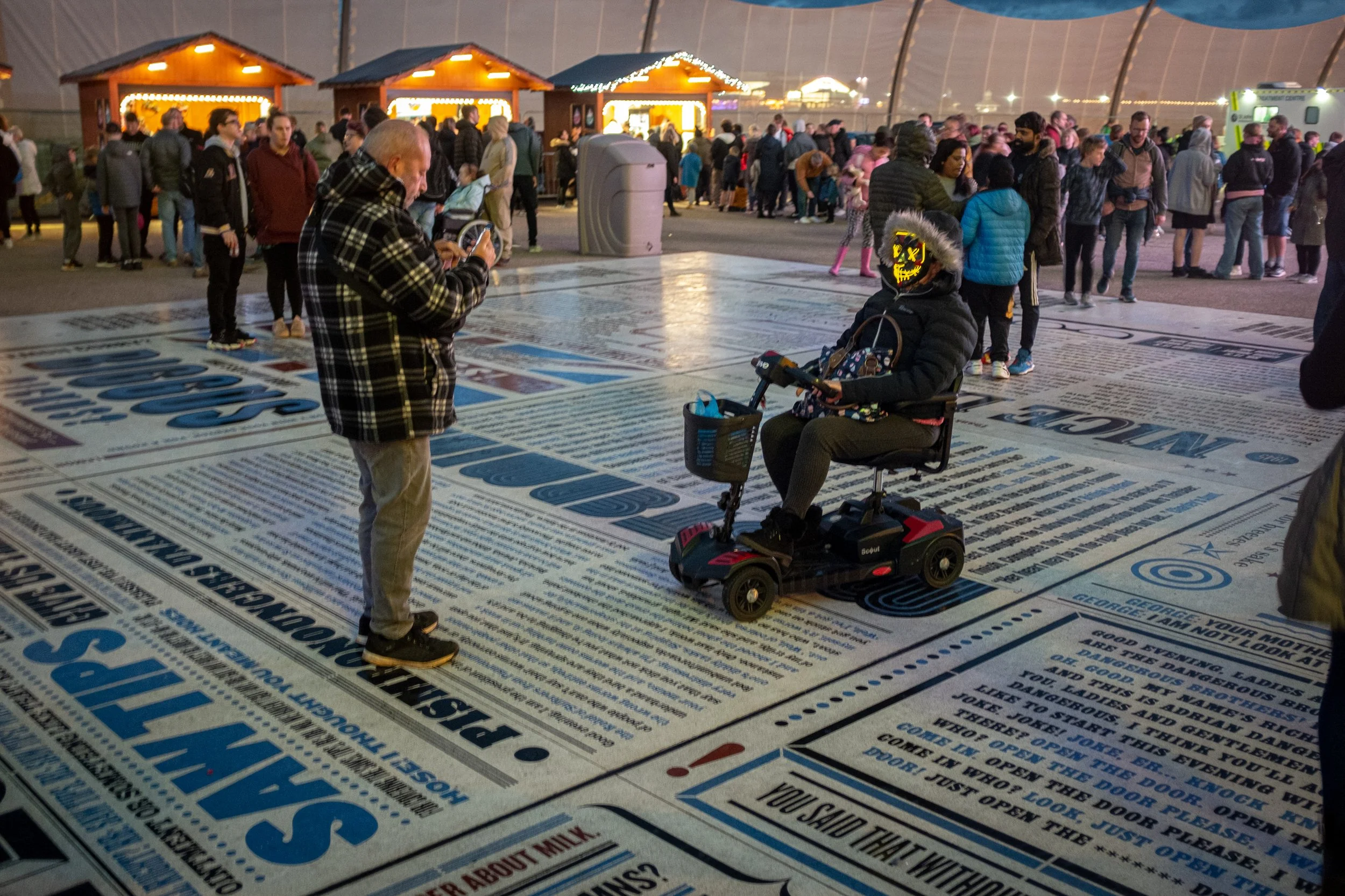People standing in line at an indoor event, with a booth in the background decorated with string lights, and a person in a wheelchair wearing a clown mask.