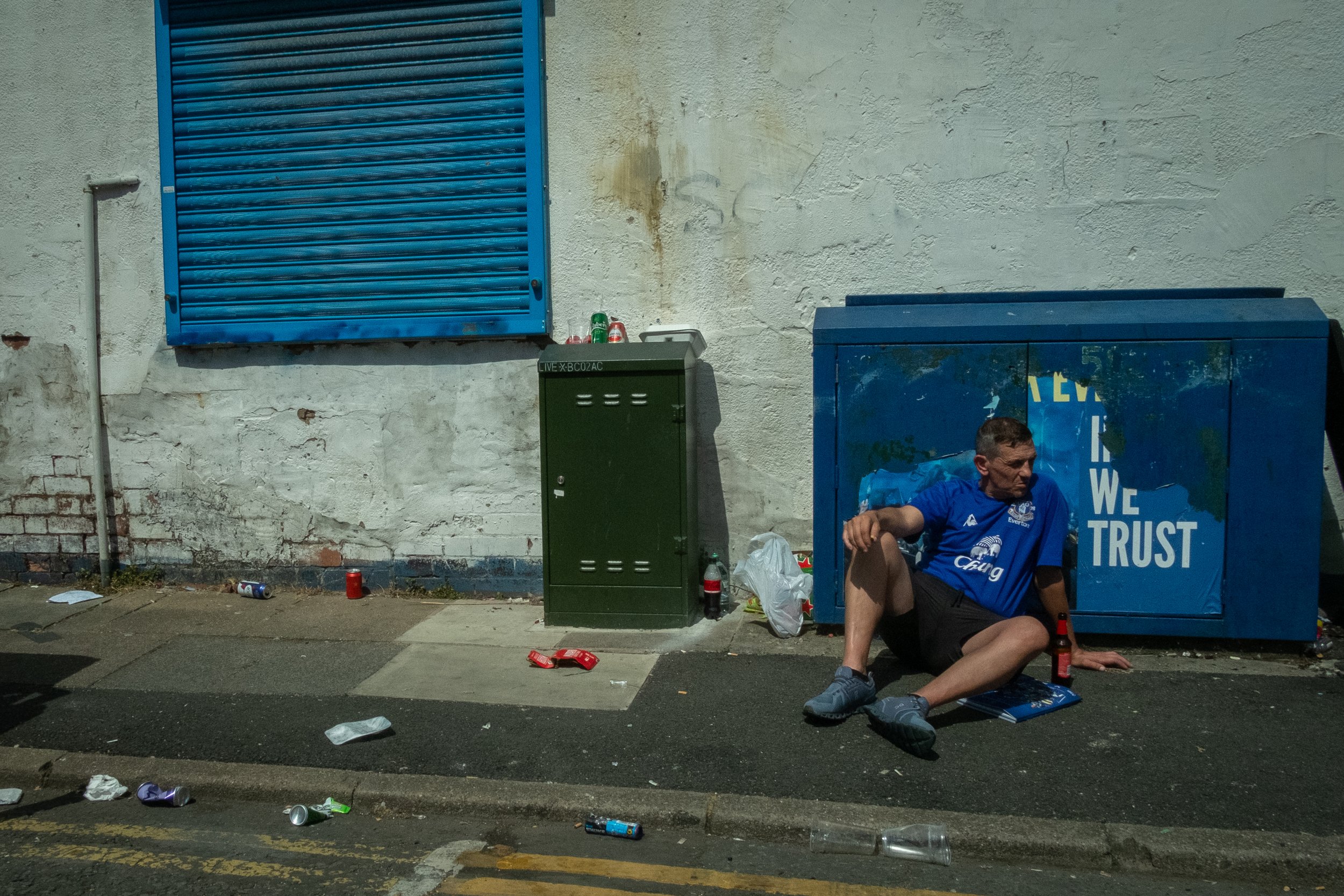 A man in a blue sports jersey sitting on the sidewalk next to a blue container with the words 'WE TRUST' on it, surrounded by litter, during nighttime.