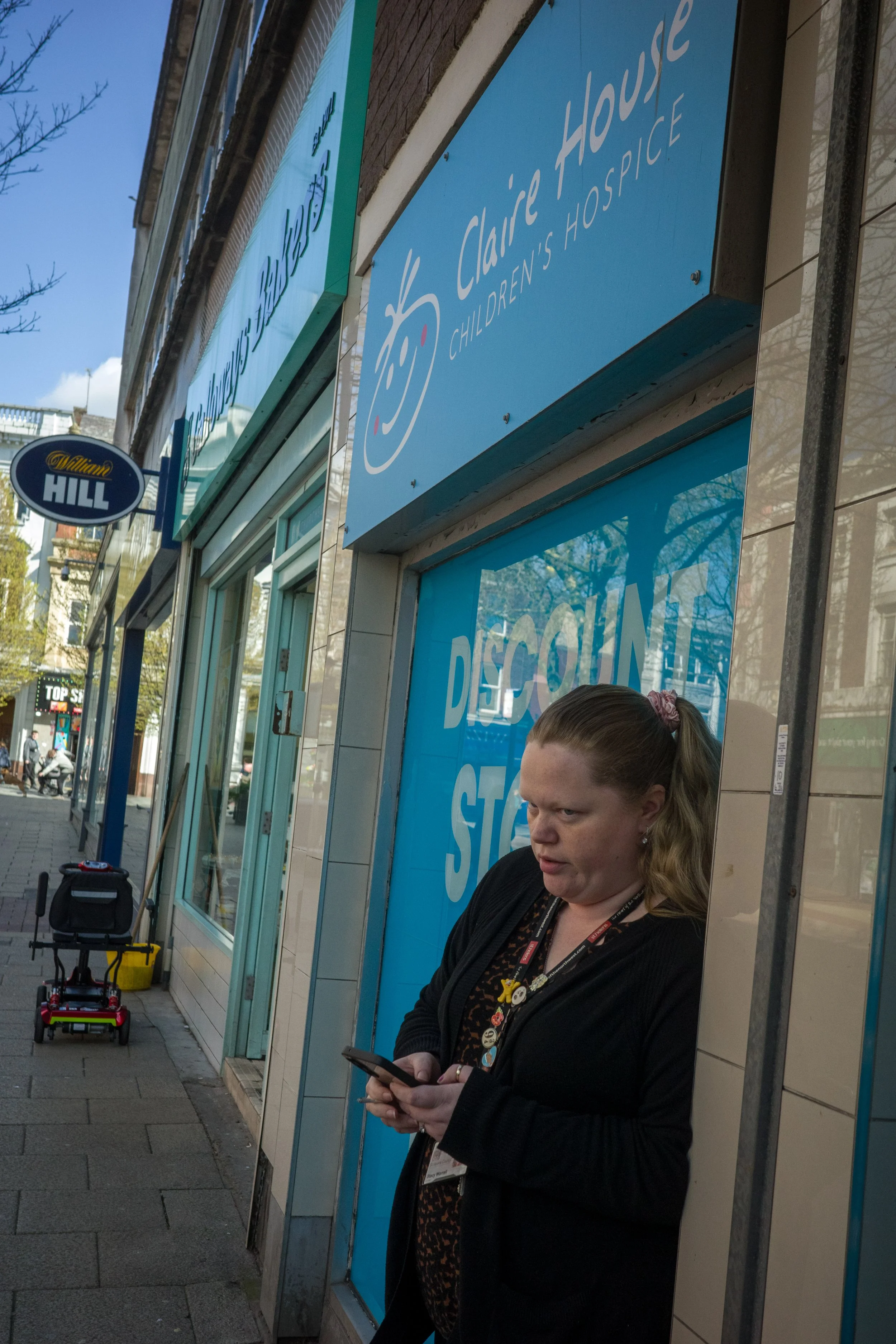 A woman with long hair tied back, wearing a black jacket and patterned shirt, is standing outside a store, looking at her phone. There are signs for Children's Claire House Children's Hospice and a discount sale on the store window. A small red and y