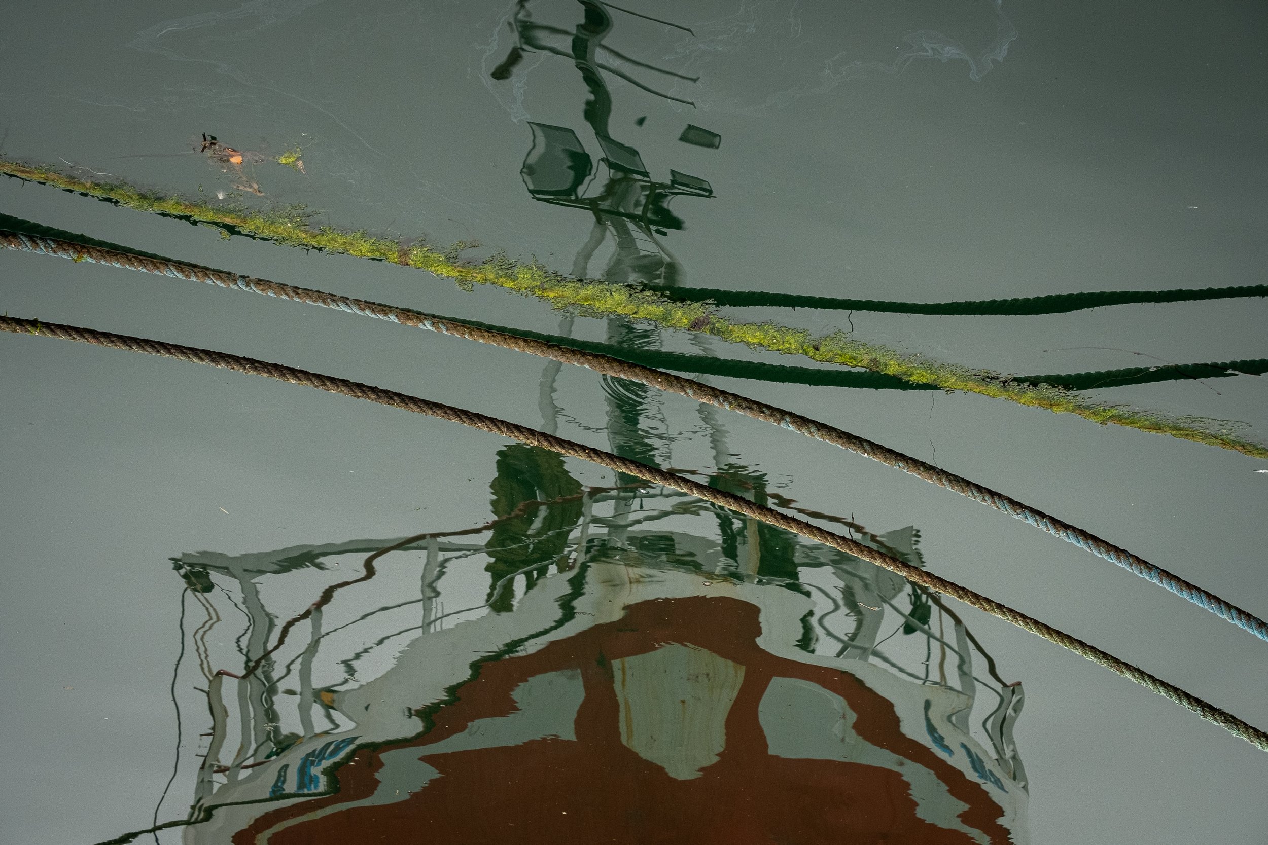 Reflected image of the bow of a boat in the water, with ropes and green algae on the water surface.