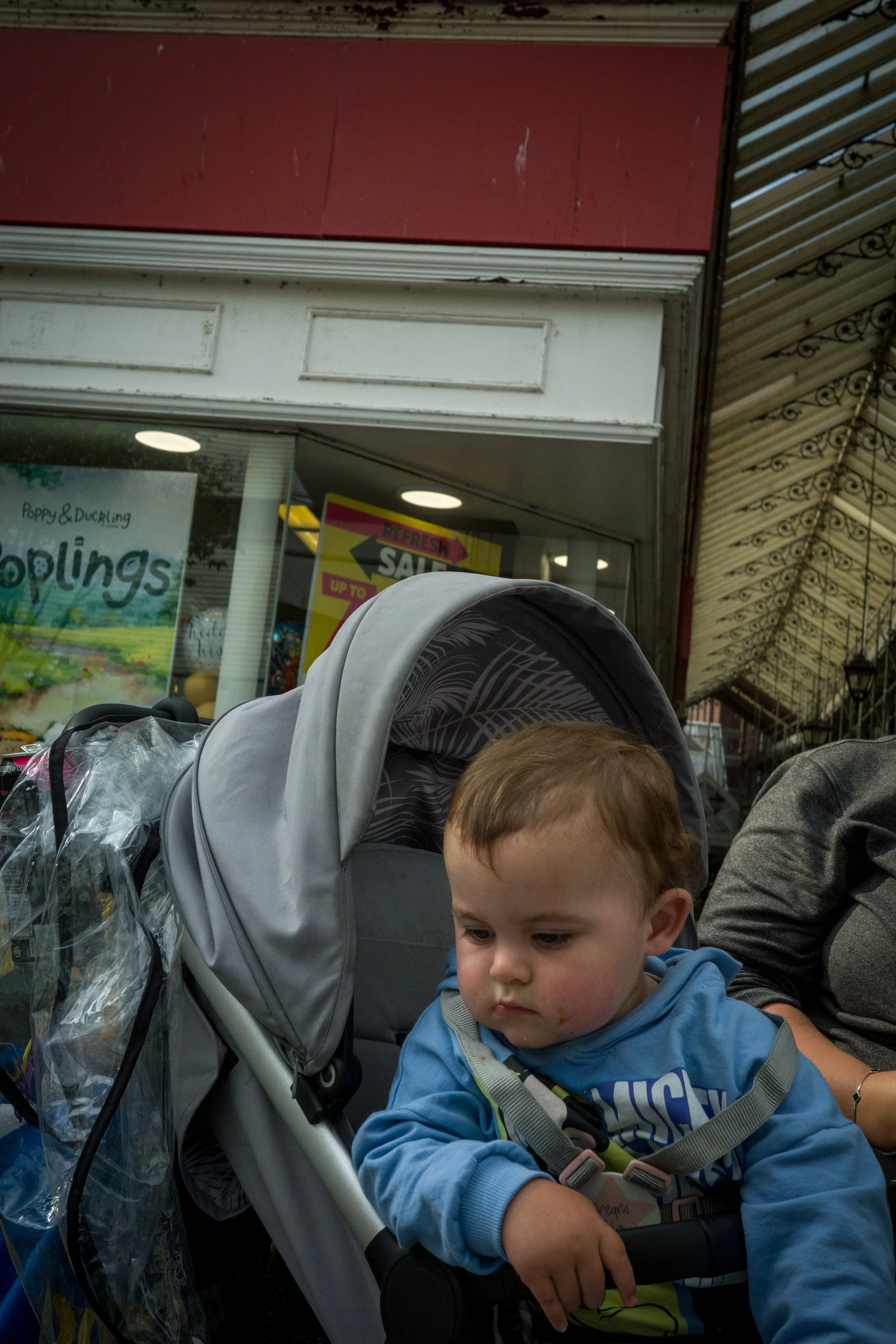 A young boy sitting in a stroller, looking down with a serious expression, outside a store with promotional signs in the background.