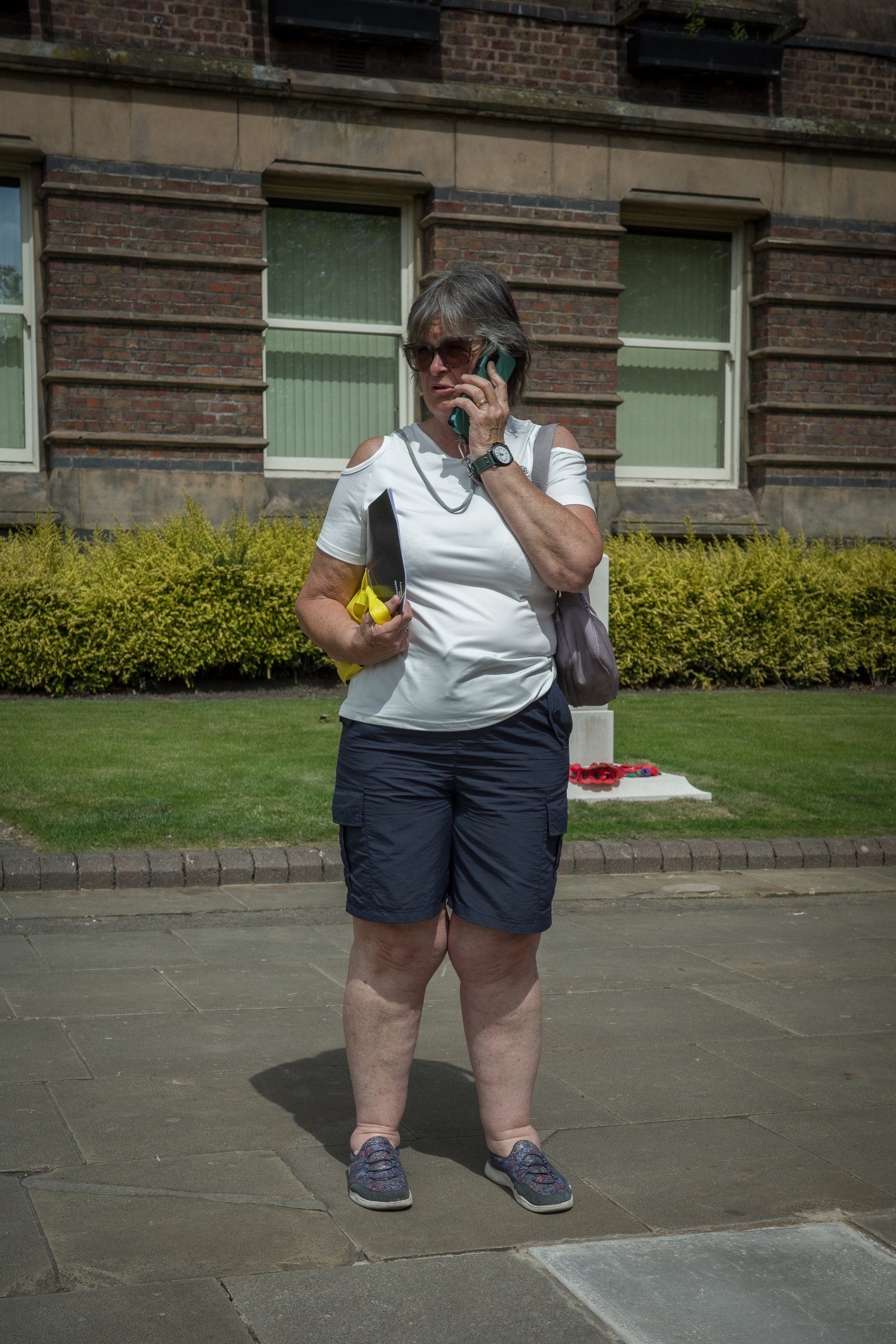 A woman standing outdoors on a sidewalk, talking on a cell phone, holding a folder, wearing sunglasses, a white top with cutout shoulders, black shorts, and sneakers, with a building and bushes in the background.