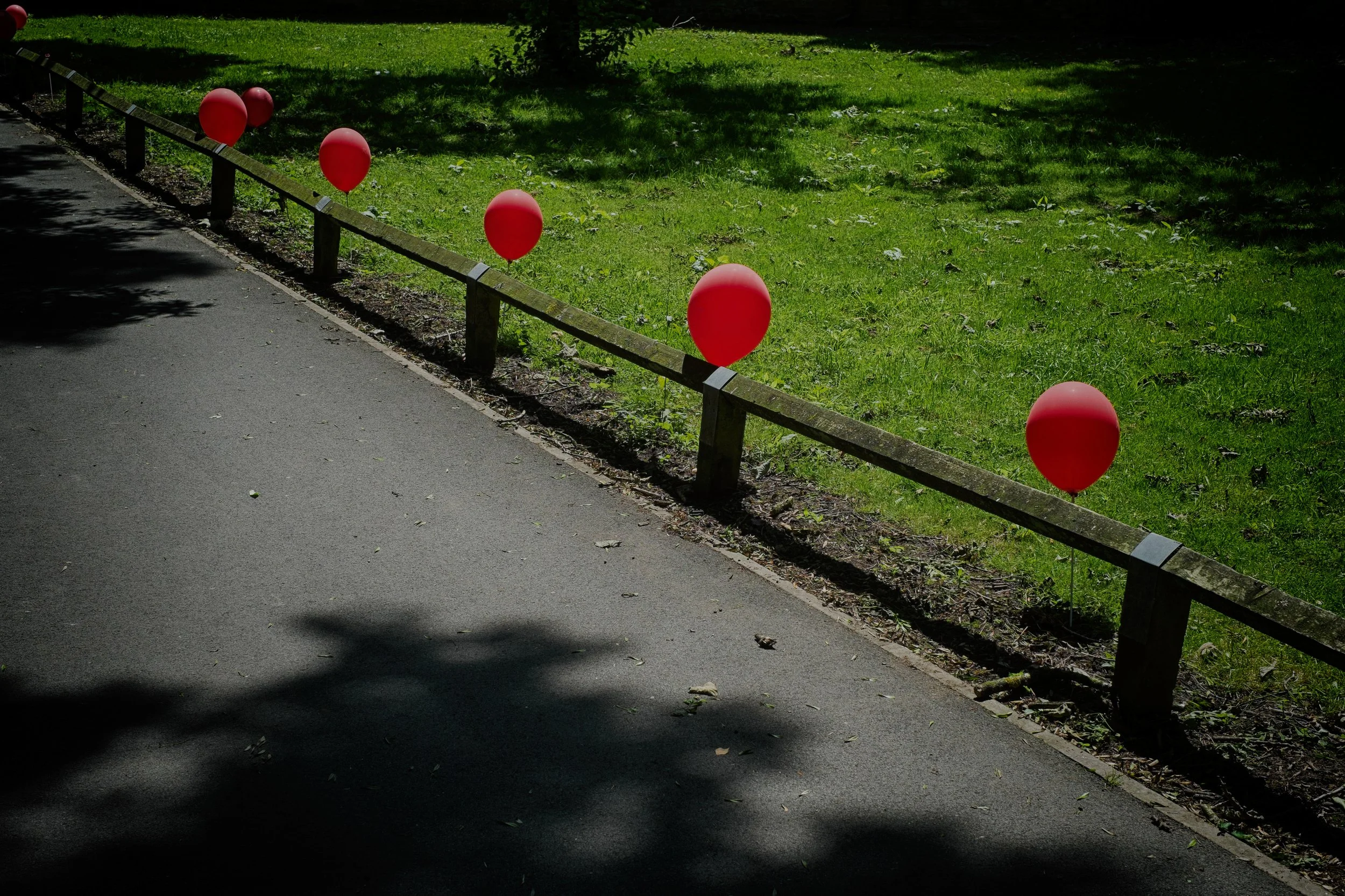 Six red balloons attached to a wooden fence along a sidewalk and grassy area, with shadows of trees on the pavement.