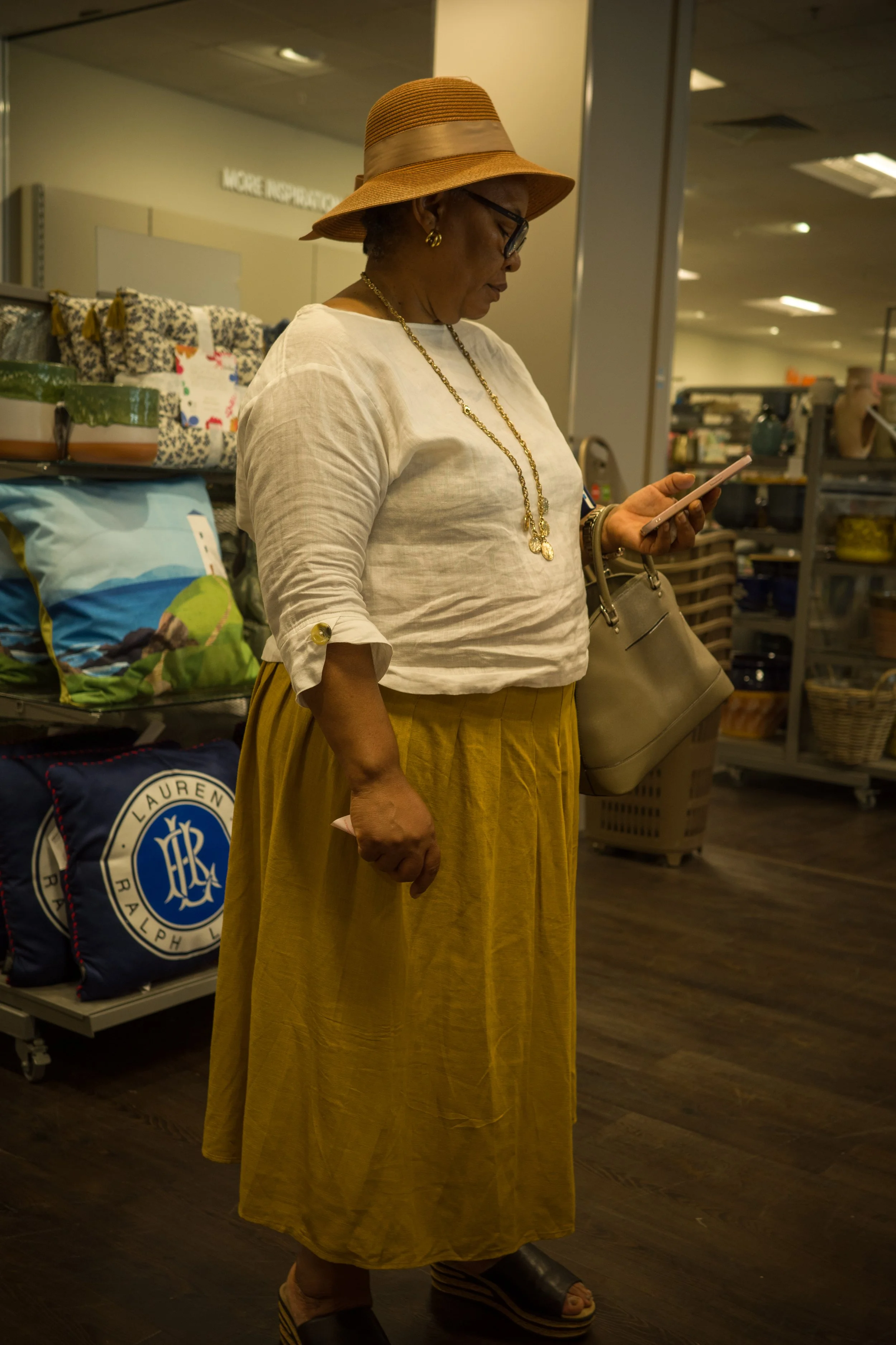 An older woman wearing a large striped sun hat, glasses, a white blouse, a yellow midi skirt, and sandals, standing indoors at a store or shopping center, holding a beige purse and looking at her phone.
