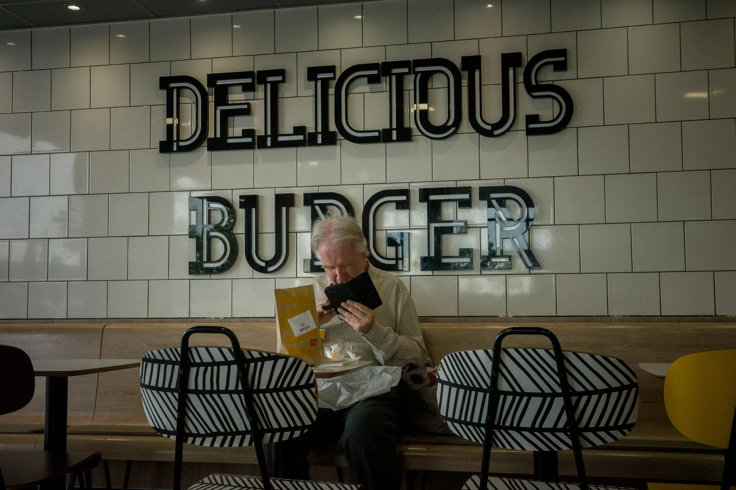 A man sitting at a table with a yellow shopping bag, looking at his phone in front of a wall with the word 'DELICIOUS' and 'BURGER' in large black metal letters.