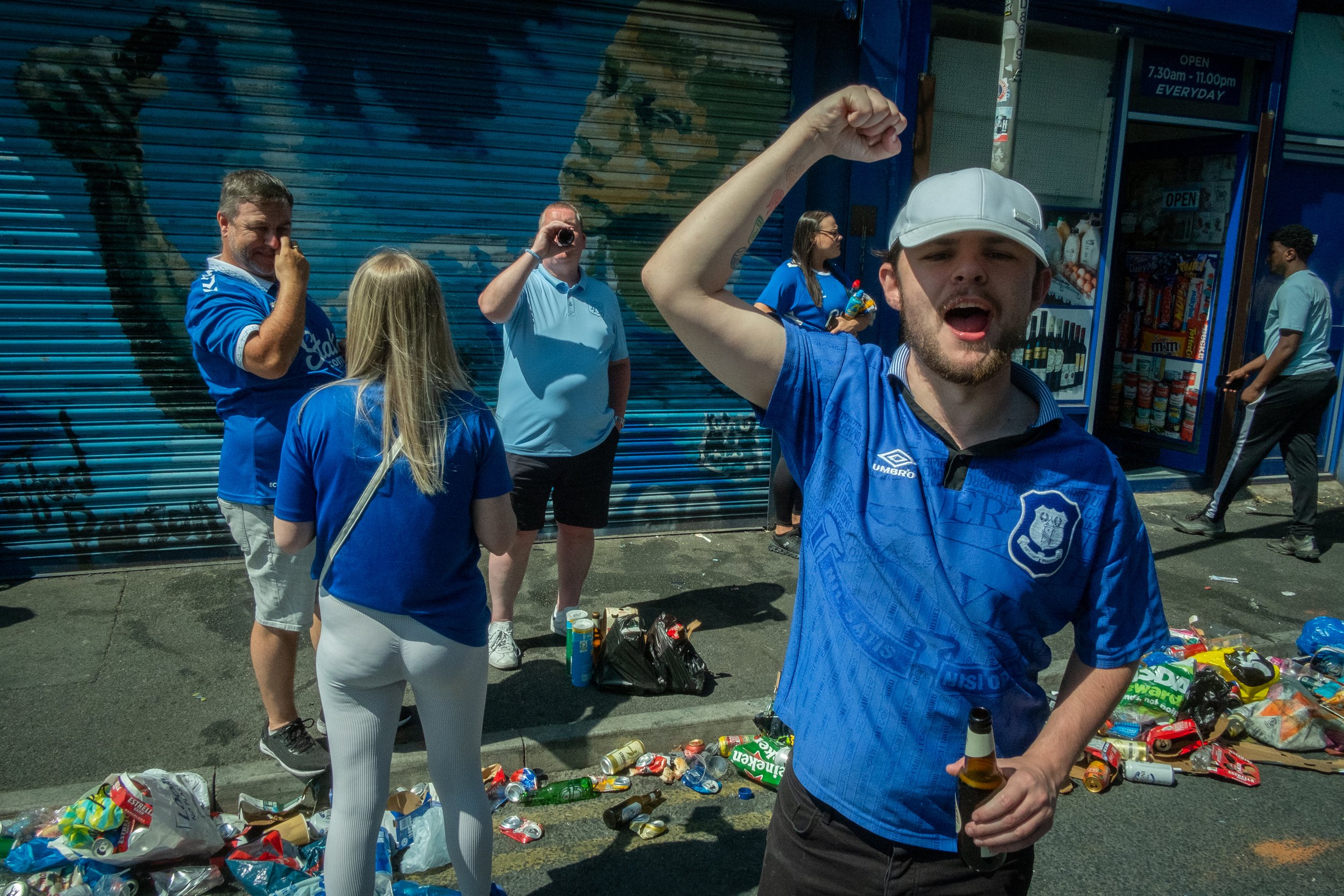 Group of people dressed in blue soccer jerseys standing and celebrating on a street sidewalk, with garbage scattered around.