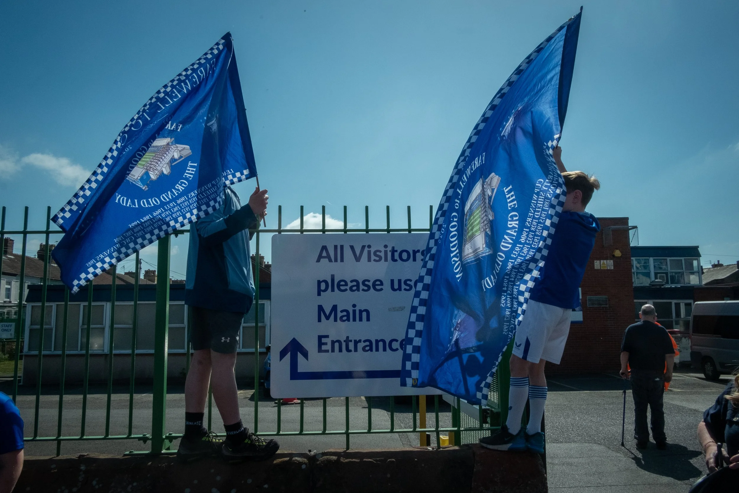 Two young men holding blue flags with a school crest near a sign that says 'All Visitors please use Main Entrance' outside a building, with other people and buildings in the background.