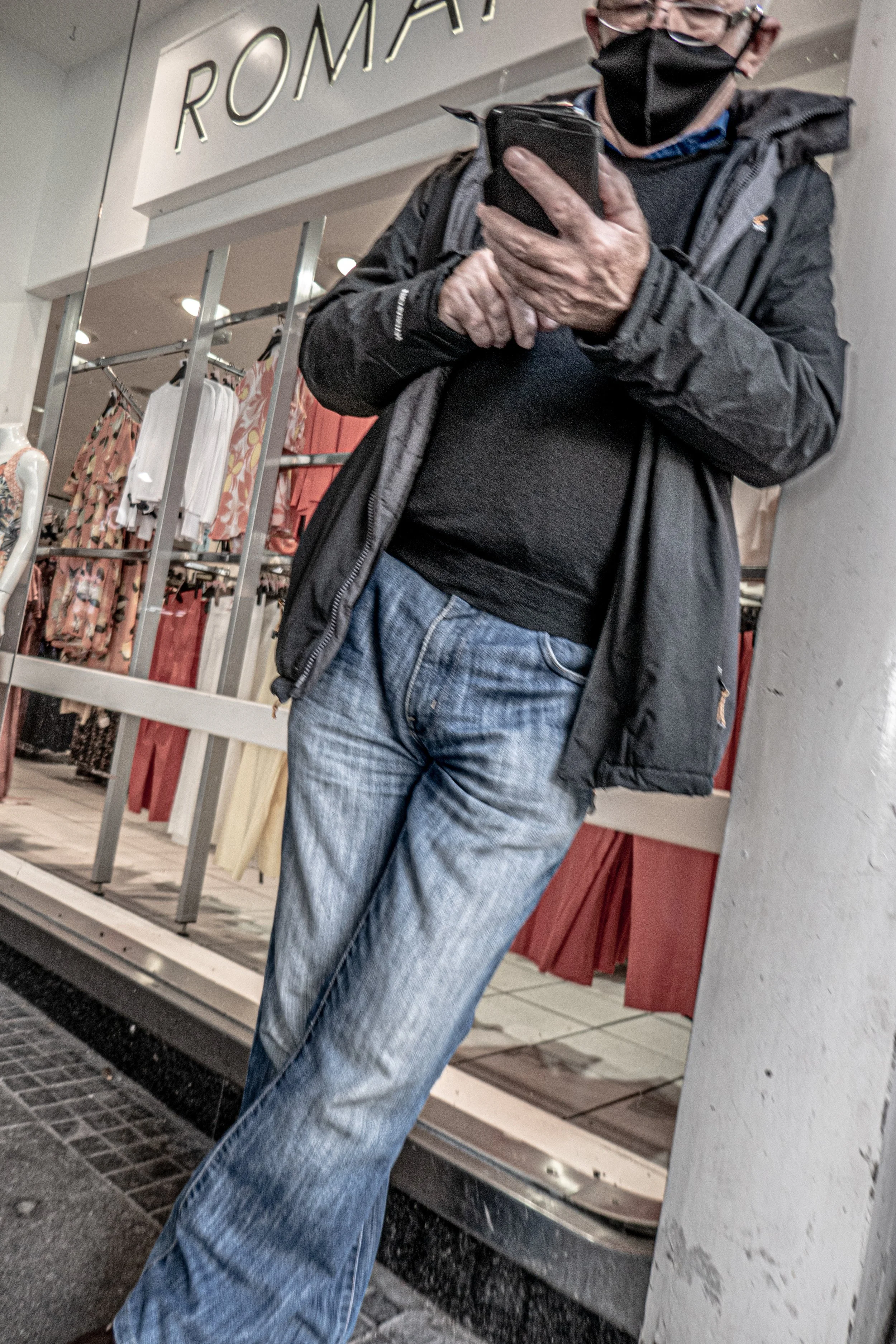 A person standing near a store window, wearing a black face mask, glasses, a black jacket, and blue jeans, looking at their phone.
