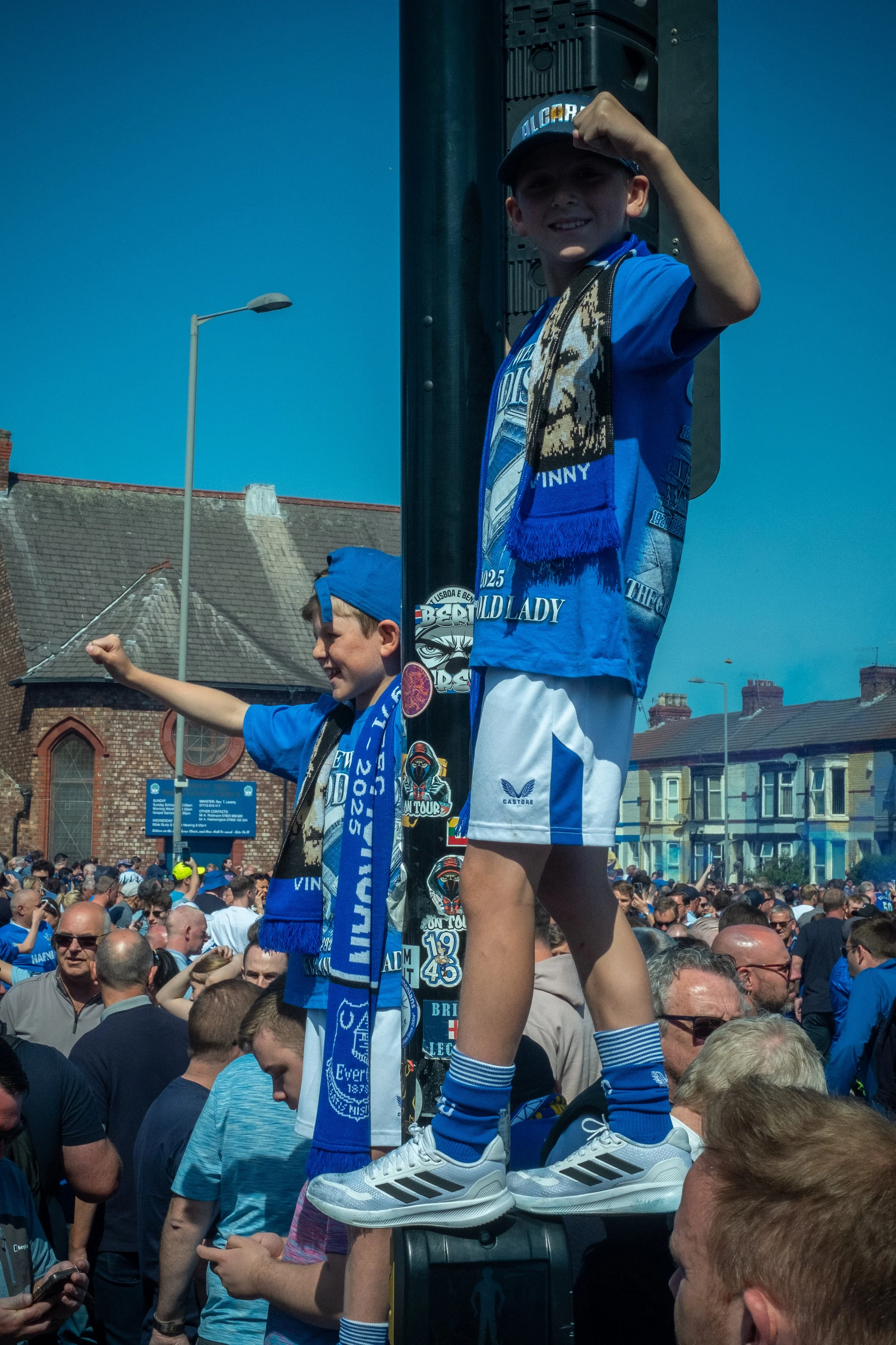 Two young boys dressed in blue and white sportswear, standing on a raised platform among a crowd of people. The older boy is wearing a baseball cap and posing with his fist raised, while the younger boy is smiling and pointing with his arm extended. 