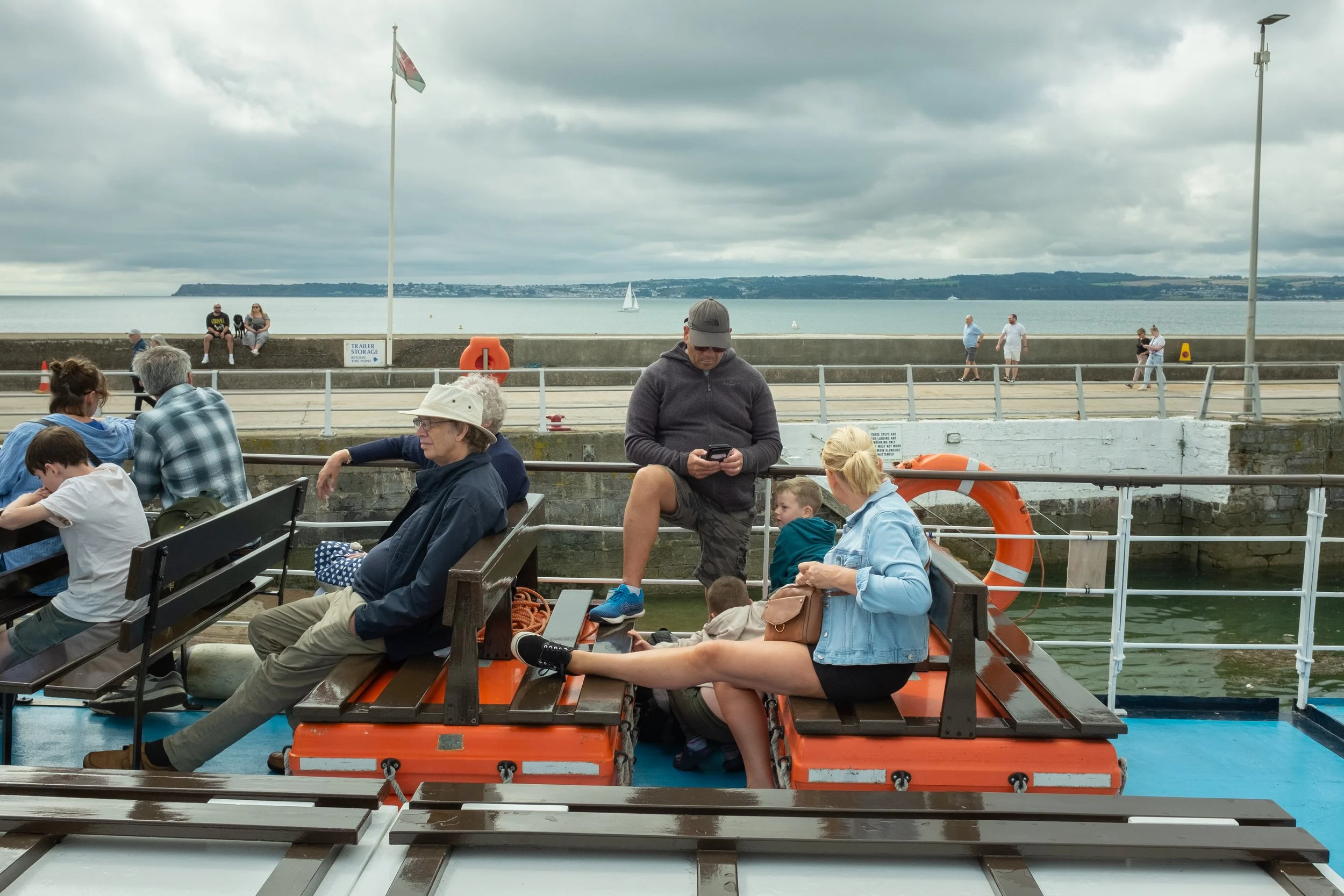 People sitting on benches and a boat in water near a port, with a cliff and sailboat in the background.