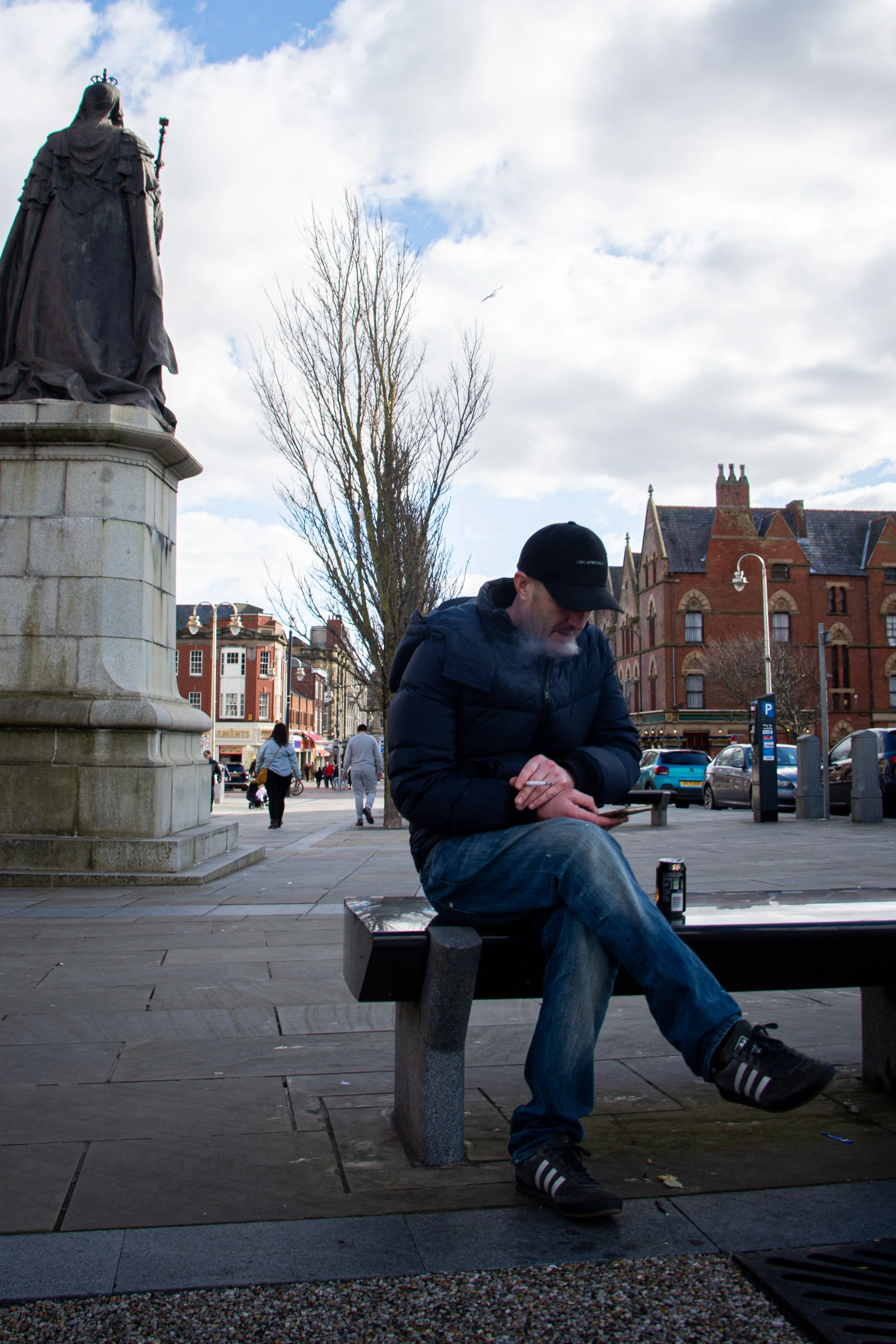 A man wearing a black cap and dark clothing sitting on a bench in a city park, using a tablet device with a can of soda on the ground beside him. There is a bronze statue on a stone pedestal of a woman with a crown and robes to his left, and building