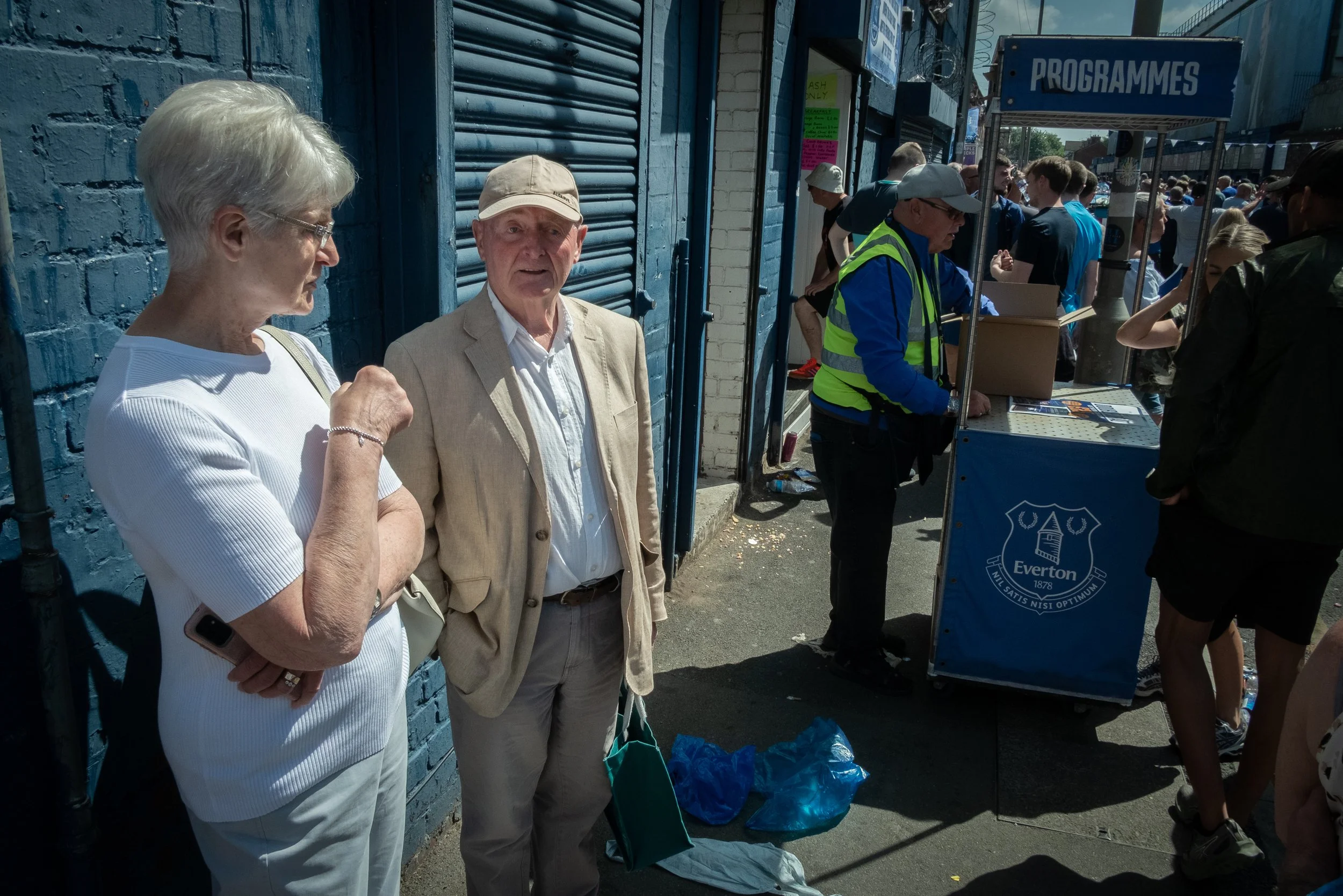 Two elderly people, a woman and a man, standing outside near a blue wall, engaged in conversation, with a crowd and a registration booth labeled 'PROGRAMMES' in the background.