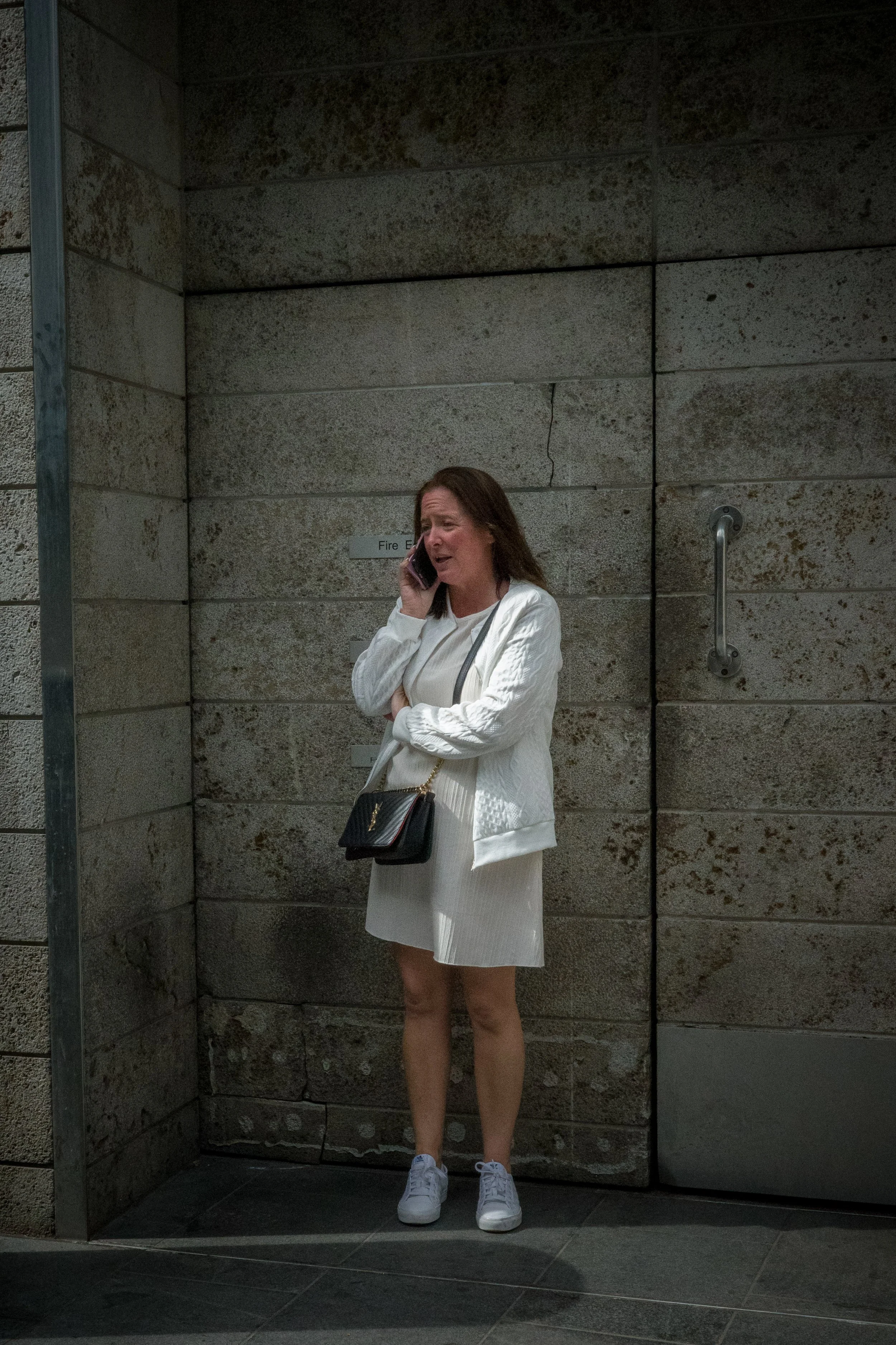 A woman with brown hair wearing a white jacket and skirt, holding a black Louis Vuitton purse, standing in front of a stone wall, talking on a cellphone.