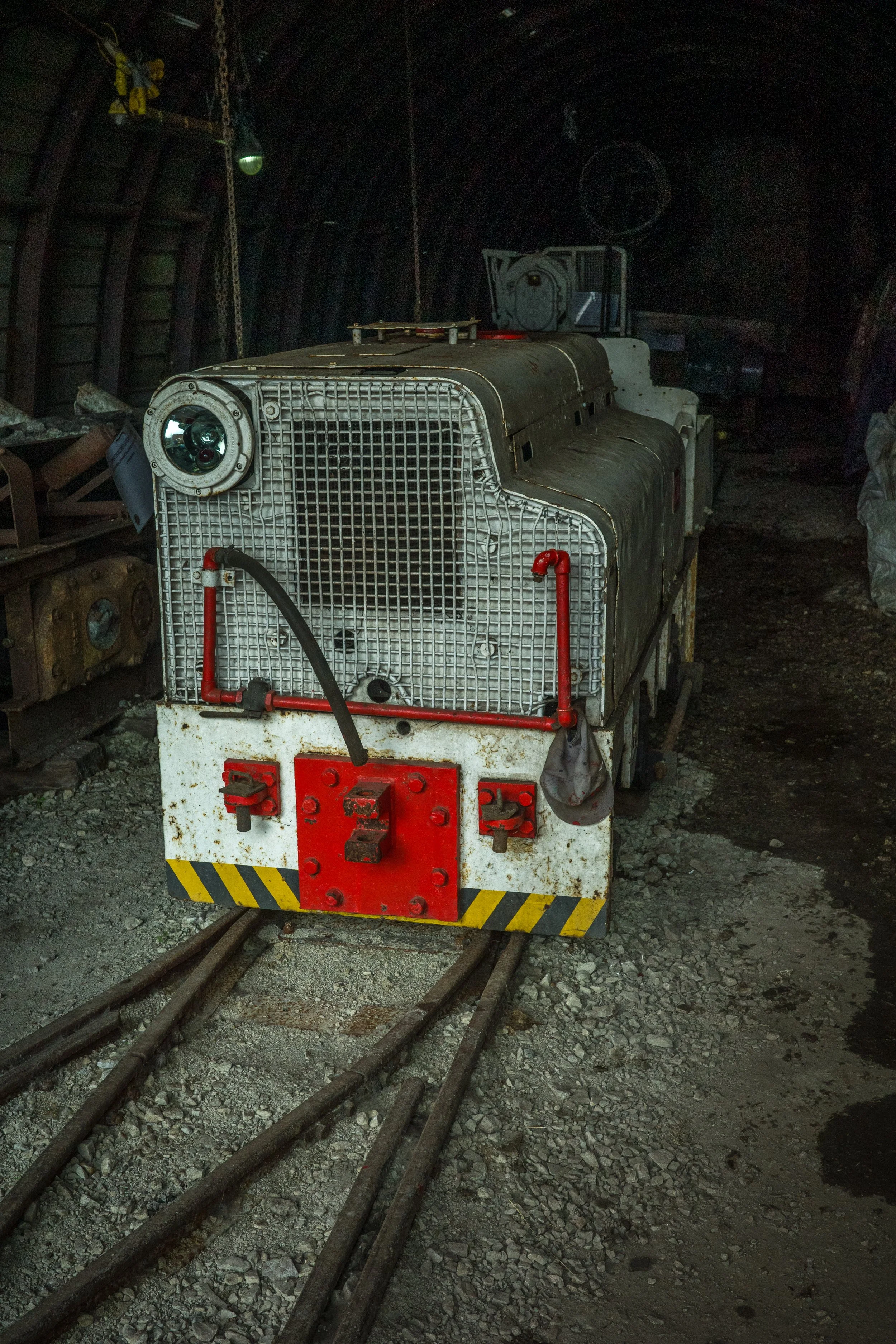A small locomotive inside a tunnel with gravel on the ground and overhead lighting, surrounded by industrial equipment and train tracks.
